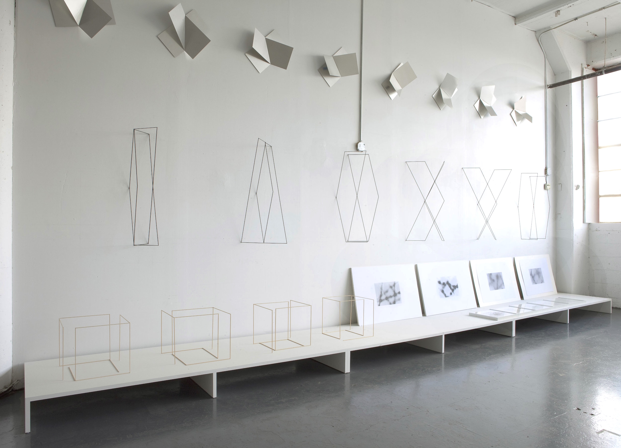 Upper wall : Untitled, 2013, 13 pairs of aluminum sheets bent at 15 ° intervals. Each pair catches the light from the window differently. Lower wall , Different But The Same, 2008, square carbon fiber extrusions, six parts, each 36 x variable x 12 in. The modular extension of a two dimensional pattern into the third dimension. Platform from left : Untitled , 2010, one-Eighth size basswood models of nine foot structures. Four drawings, 2013, Drawing As Photography ( a remembrance ), graphite and acrylic fixative on paper. Photograph: David Allison 06-14.