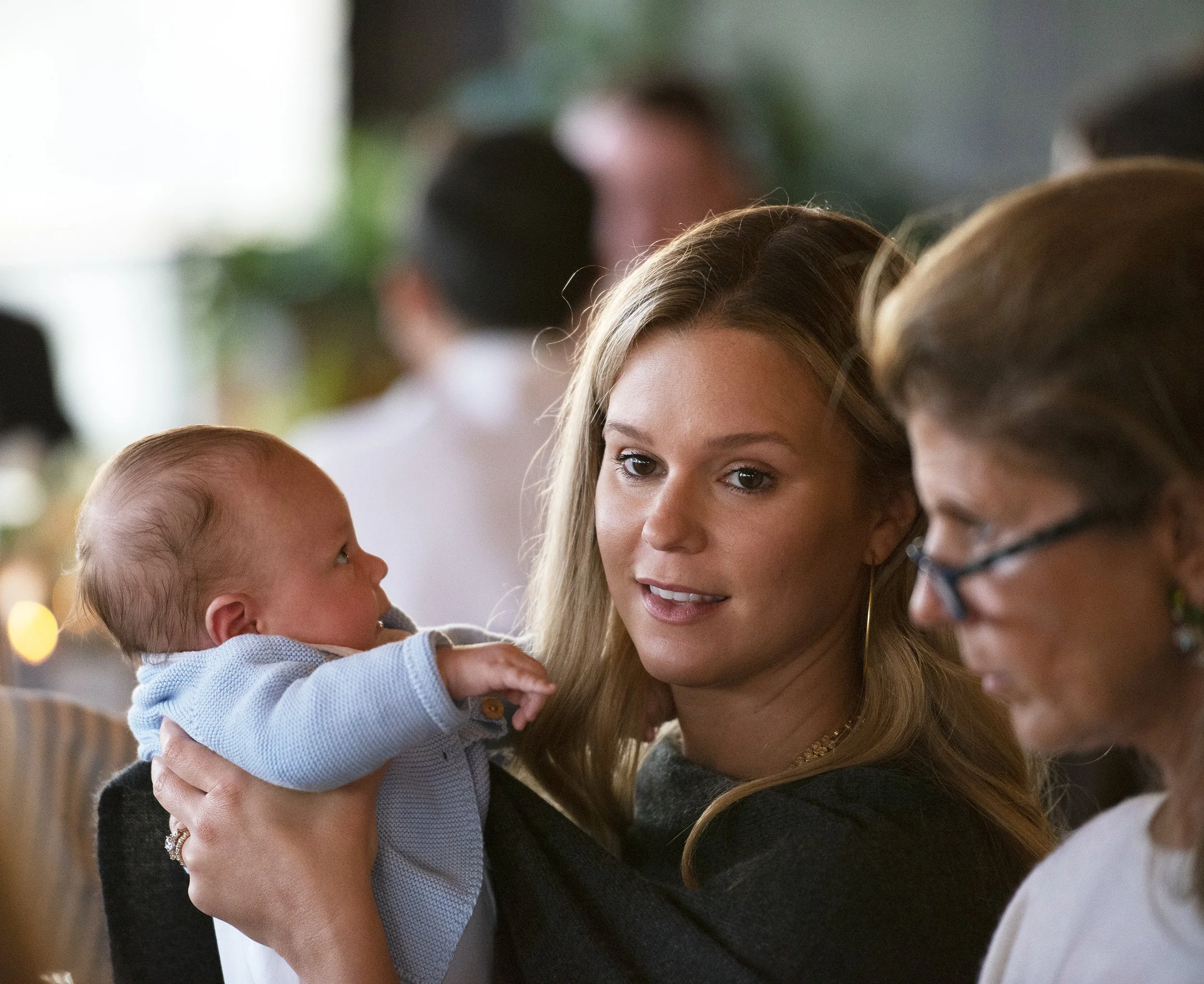 A woman with long blonde hair holding a baby in a blue shirt, engaging with two young children at a social event.