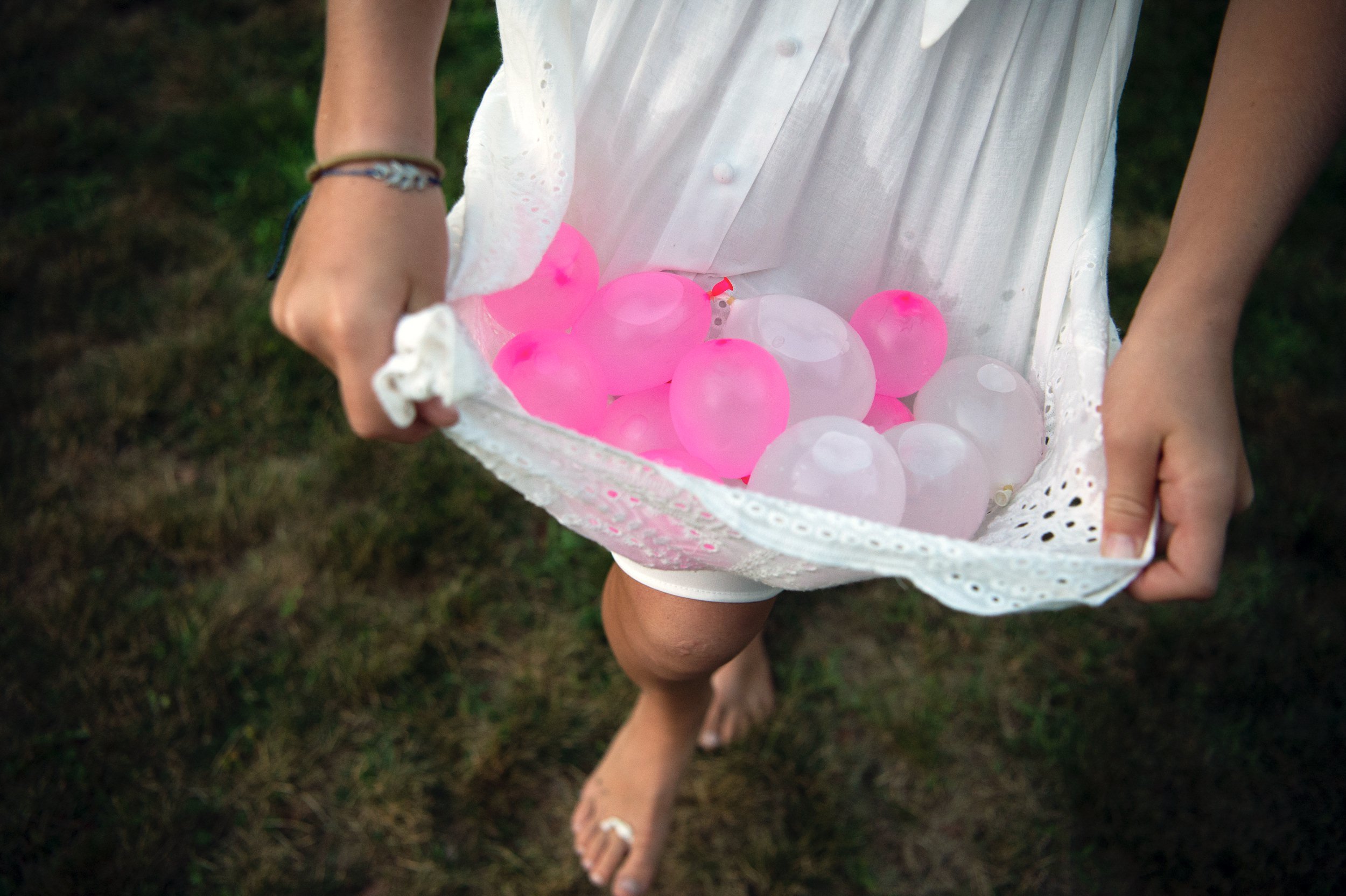 Child holding water balloons, walking barefoot on grass.