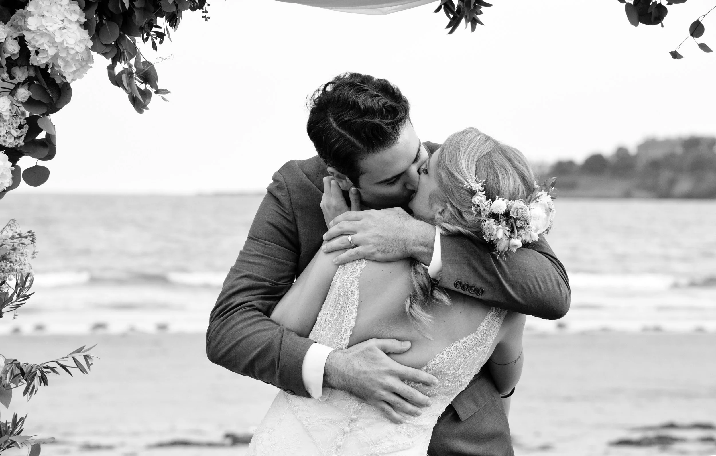 Black and white of bride and groom embracing after the ceremony with sand and ocean in background, with hair by Studio B Salon
