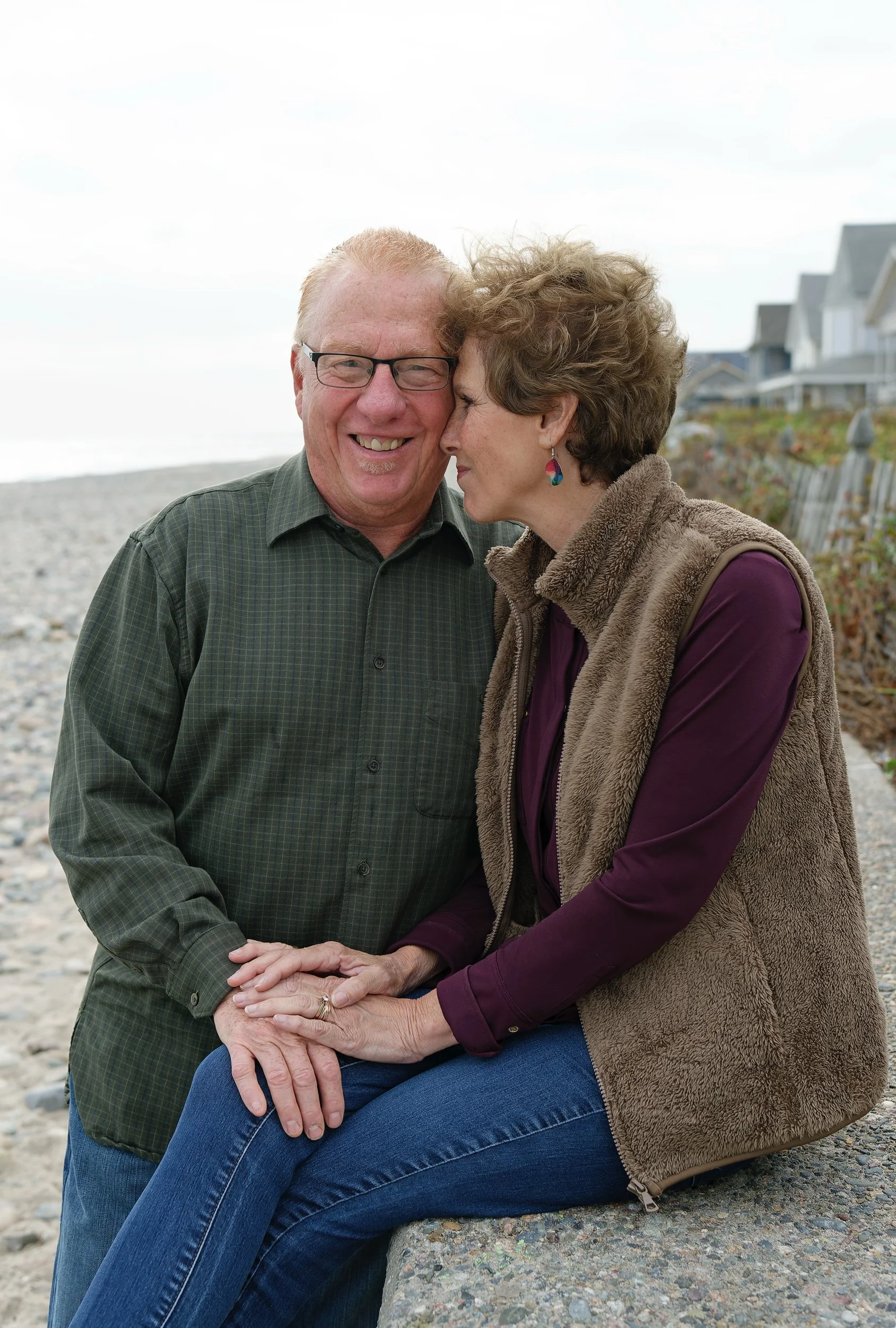 A man and woman sitting on a pebbled beach, smiling and holding hands, with a row of beach houses in the background. South Shore, Massachusetts.