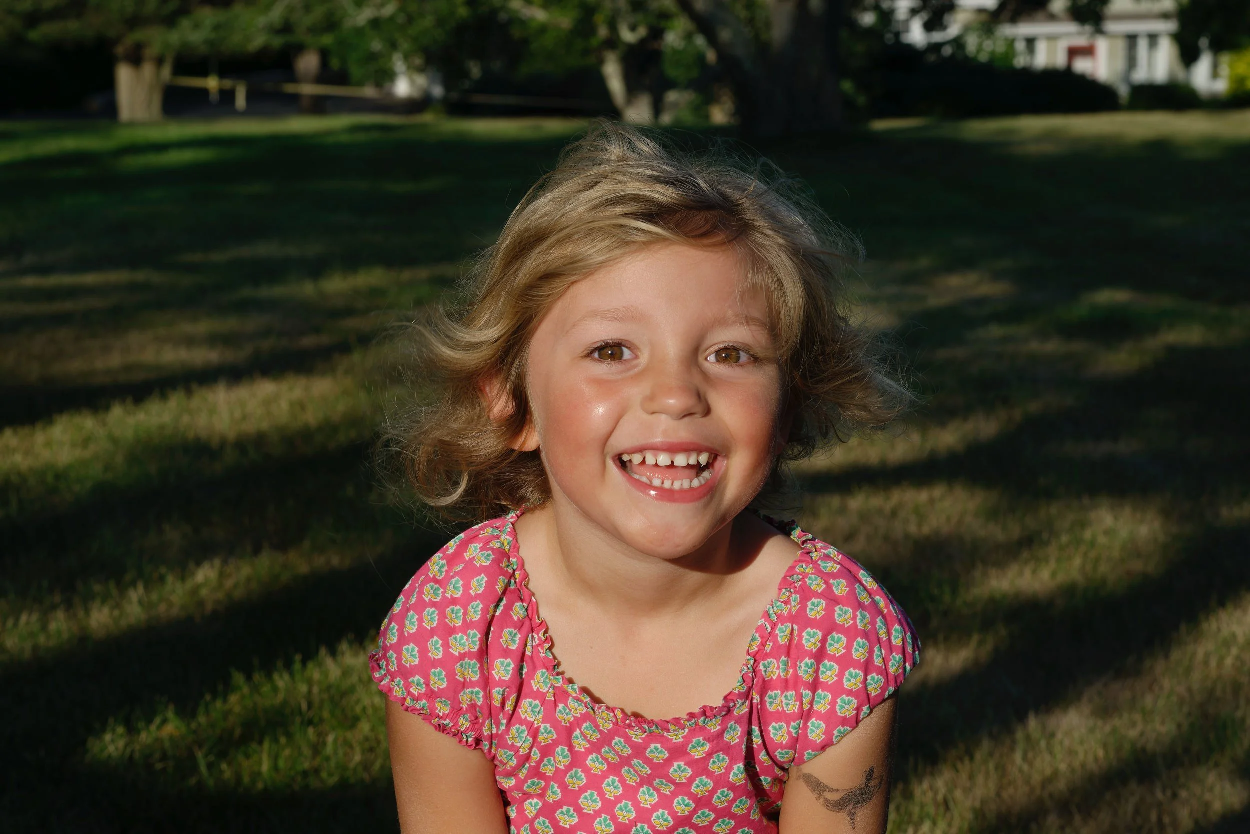 A young girl with curly blonde hair smiling outdoors in a park during the golden hour, wearing a pink floral dress.