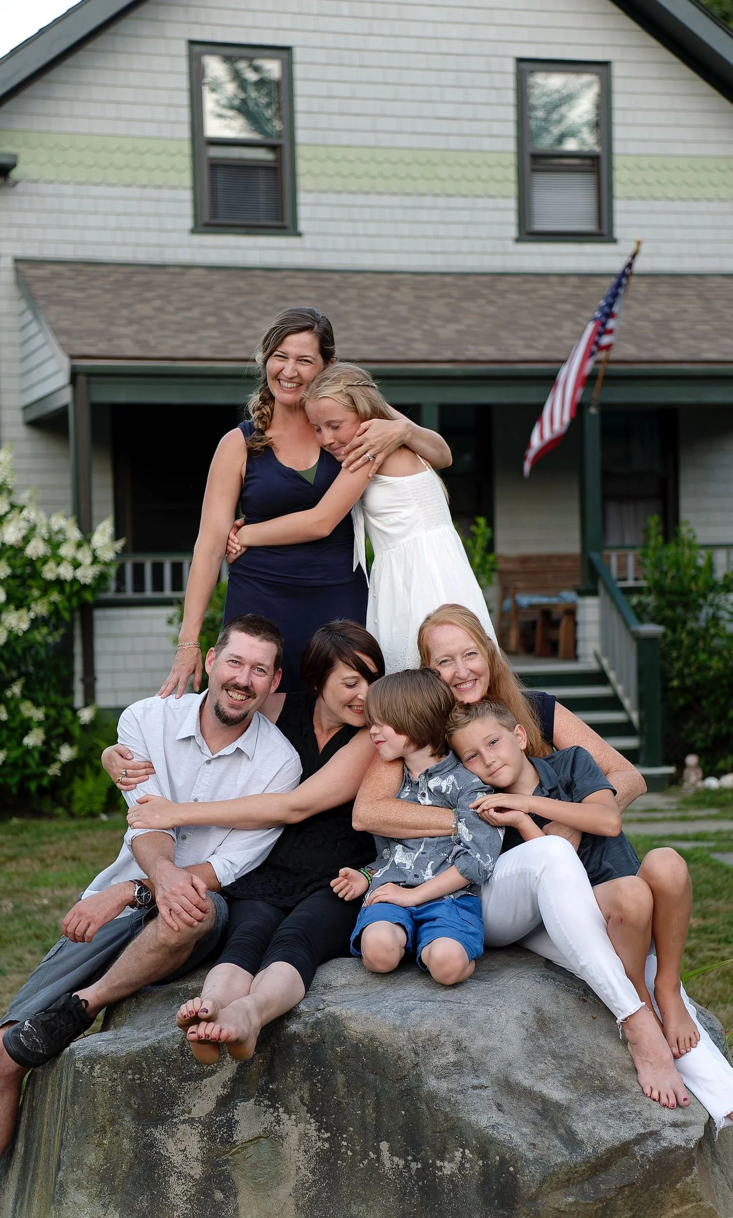 Group photo of cousins in front of a house. Seven people are sitting and standing on a large rock, smiling and embracing each other. The house has a porch, two windows, and an American flag. Jamestown, RI.