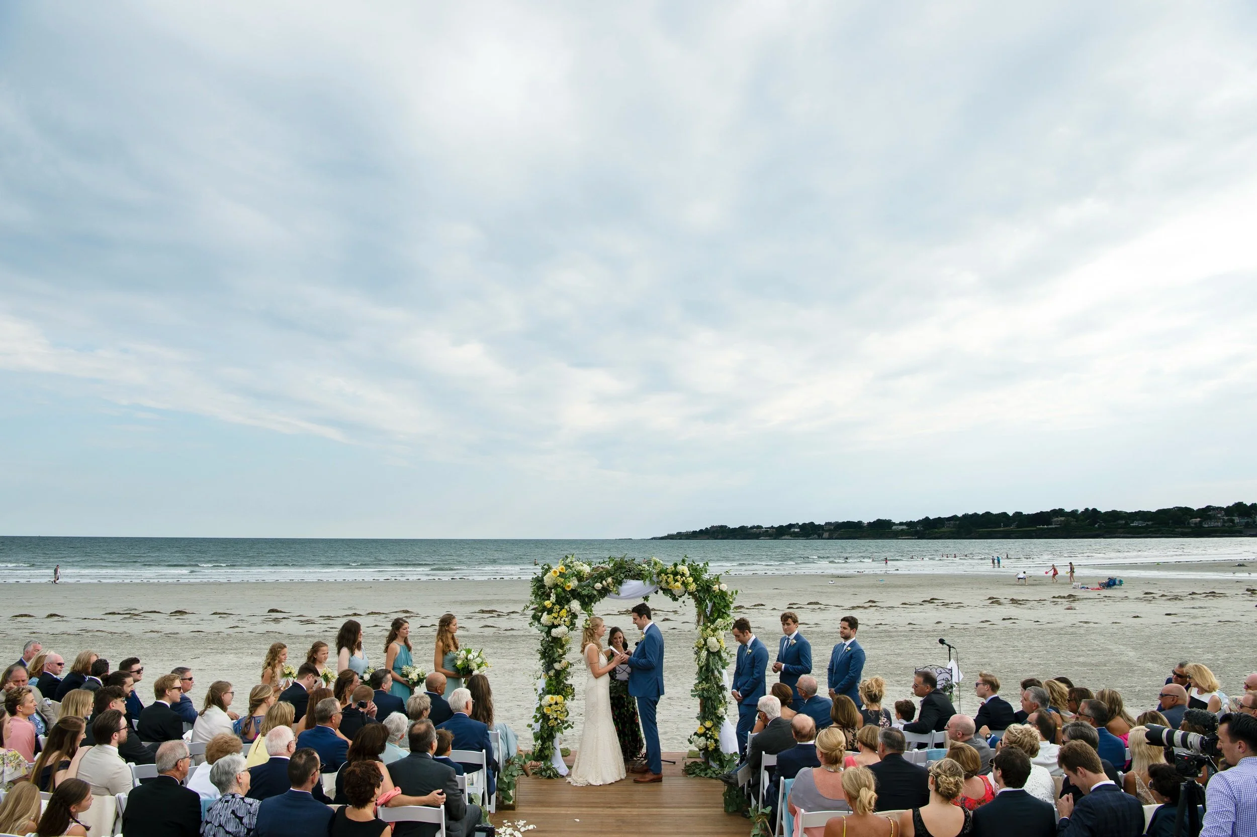 Wide-angle of bride and groom and guests during their outdoor wedding ceremony on the beach of Newport Beach House, planning by Exquisite Events and florals by Stoneblossom 