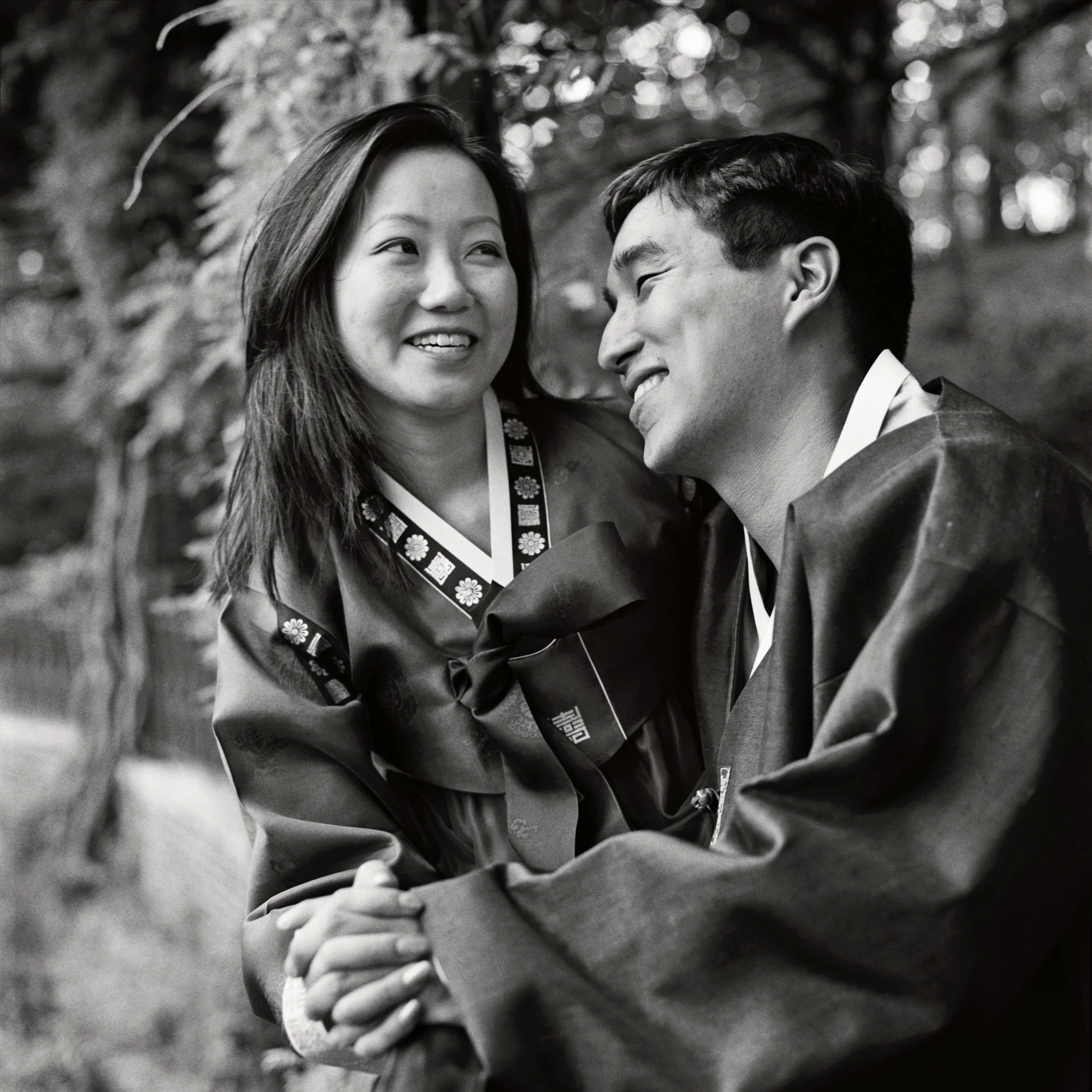 A black and white photo of a man and woman smiling and looking at each other outdoors. The woman has long hair and is wearing a floral patterned shirt, while the man has short hair, is dressed in a suit, and is leaning towards her with a smile. They 