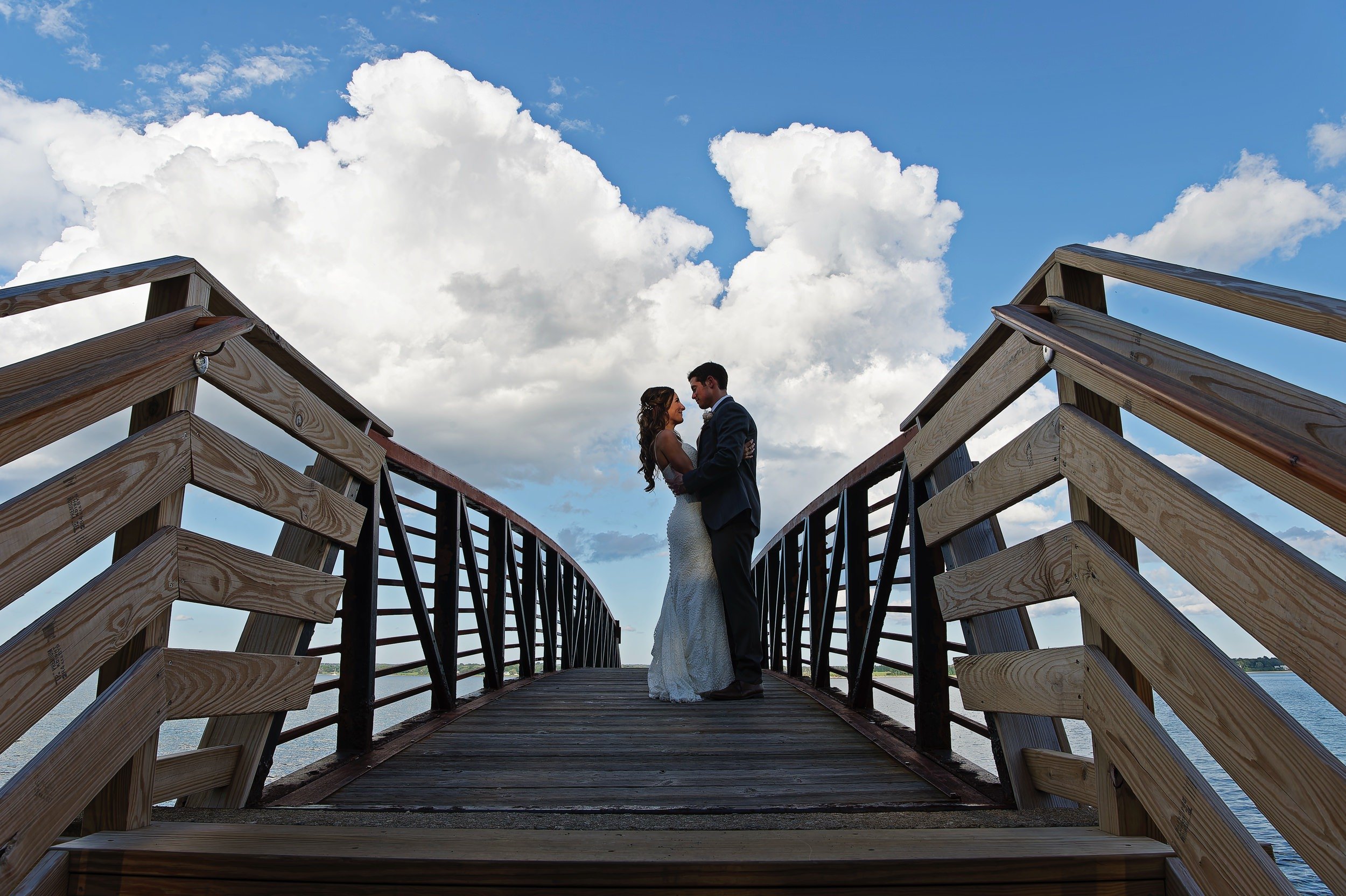 A bride and groom standing close together on a wooden bridge under a partly cloudy sky at Glen Manor in Portsmouth, RI.