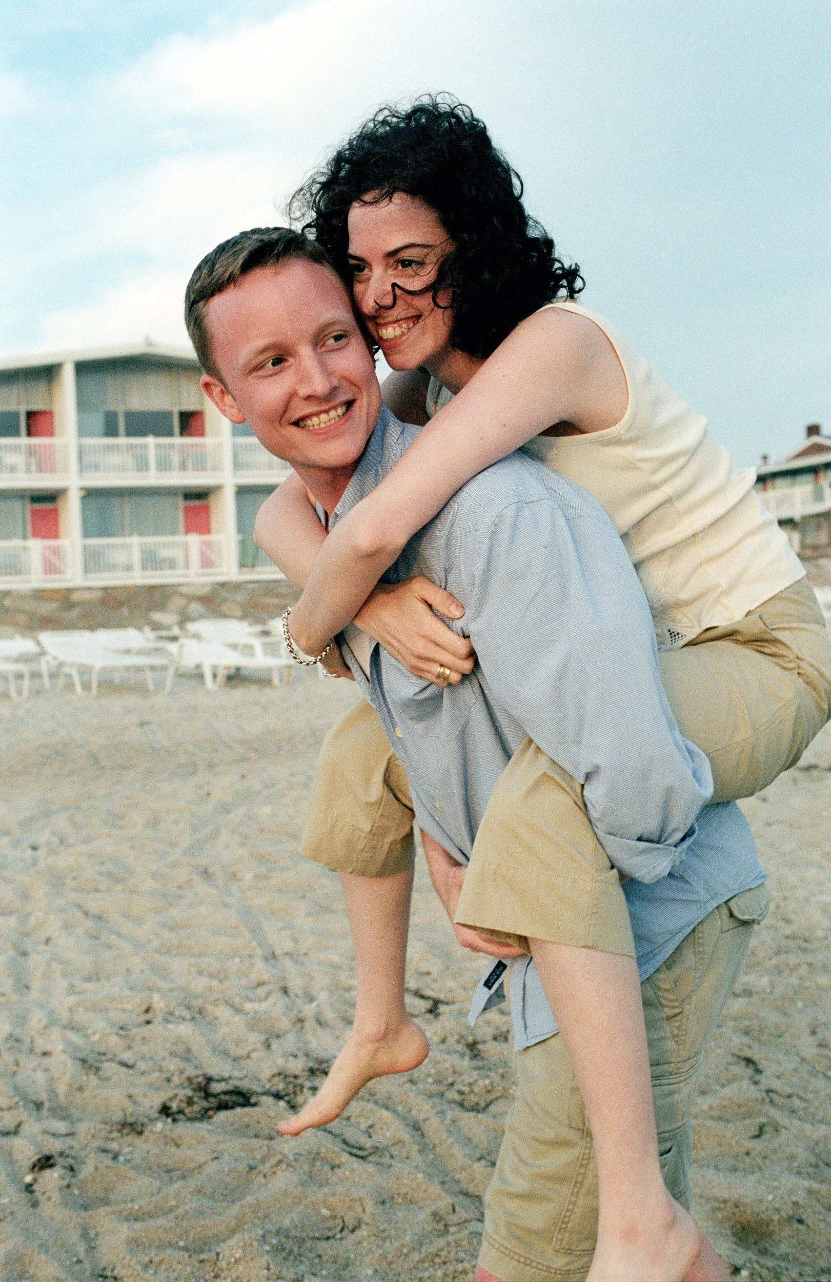 A young man giving a piggyback ride to a woman on a sandy beach with beach chairs and a modern motel or hotel in the background. North Shore, Massachusetts.