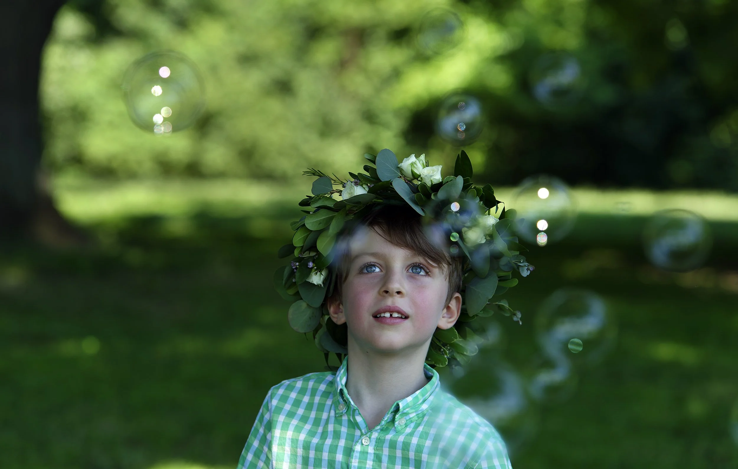 A boy blowing bubbles during a family session in Brooklyn, NY. His expression is pure joy and full of wonder as he watches the bubbles float around him. The color is very rich, full of deep and bright greenery of the surrounding nature.