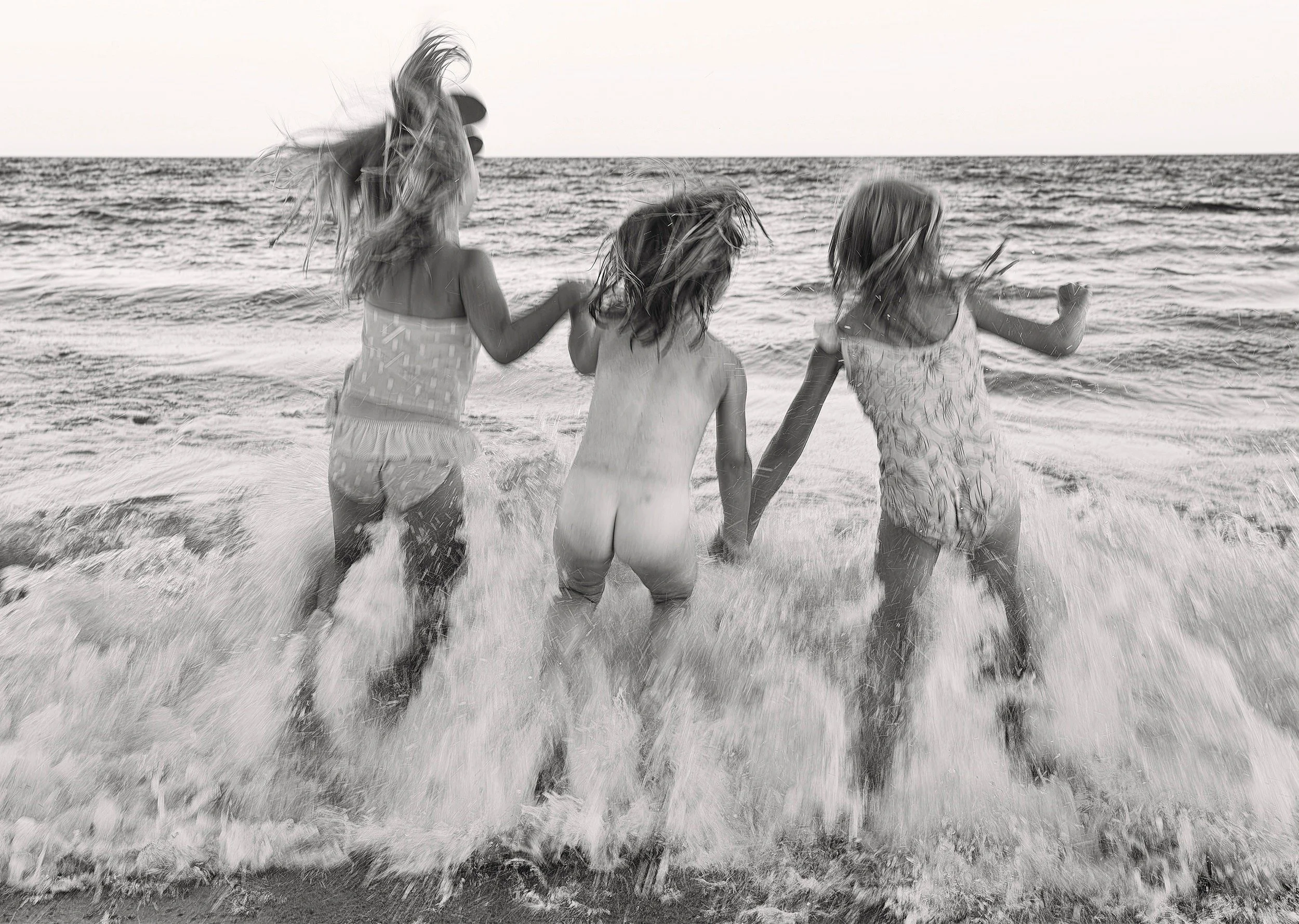 Three sisters playing at the beach with waves splashing, captured in black and white.