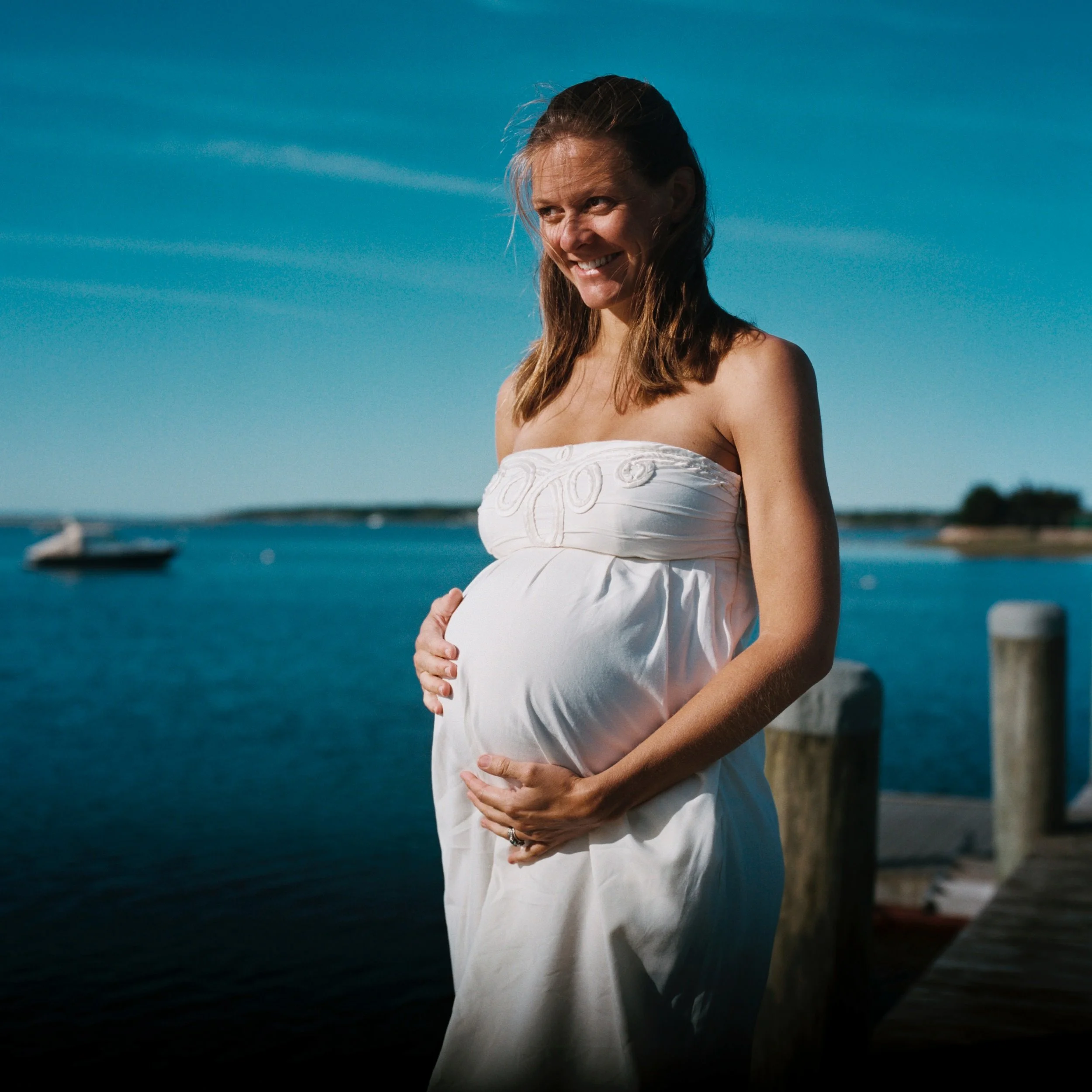 A pregnant woman wearing a white strapless dress is standing outdoors near a body of water, smiling while holding her pregnant belly with both hands. The sky is blue with a few clouds.