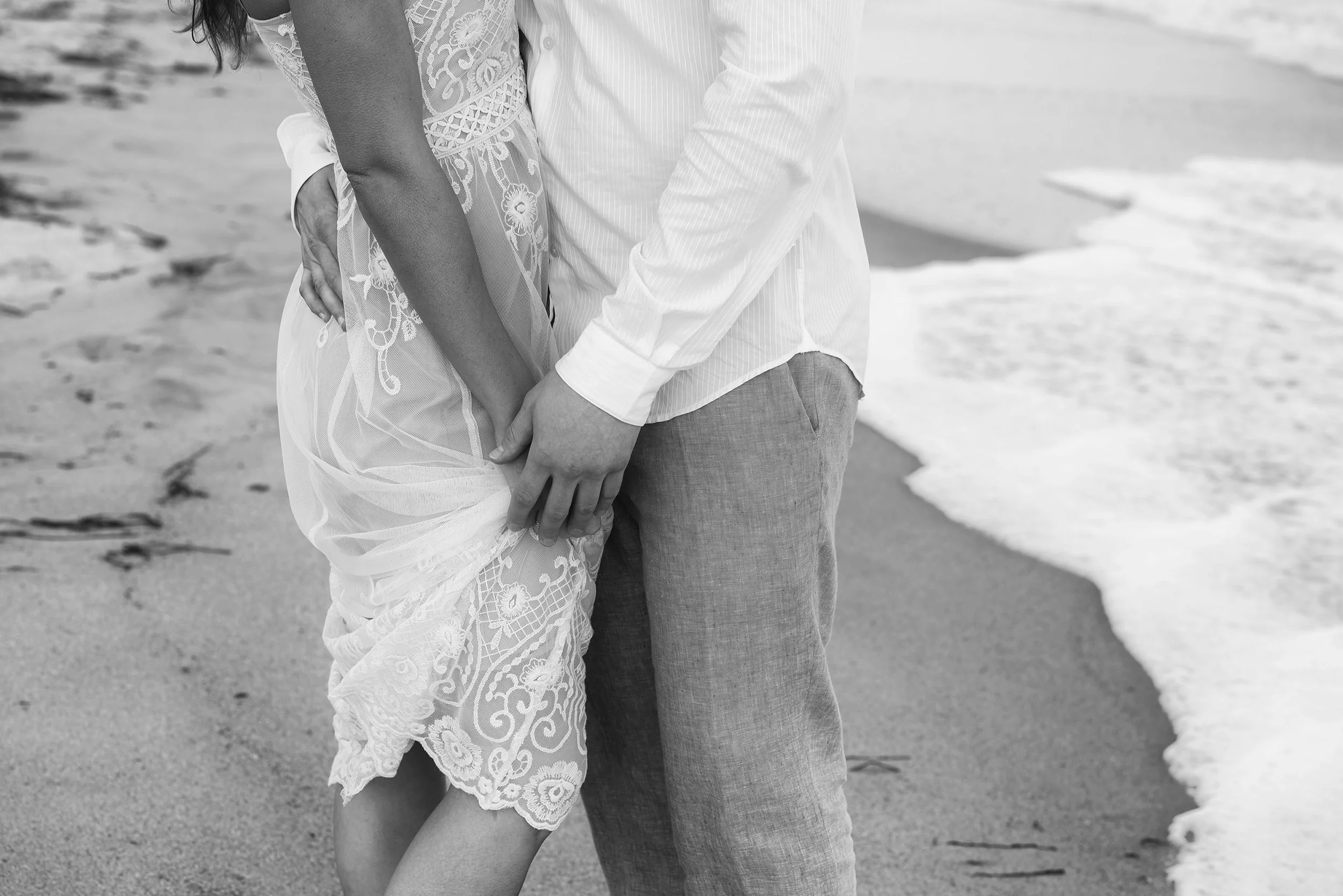 A couple holding hands on the beach, close-up of their midsections, with waves in the background, in black and white.