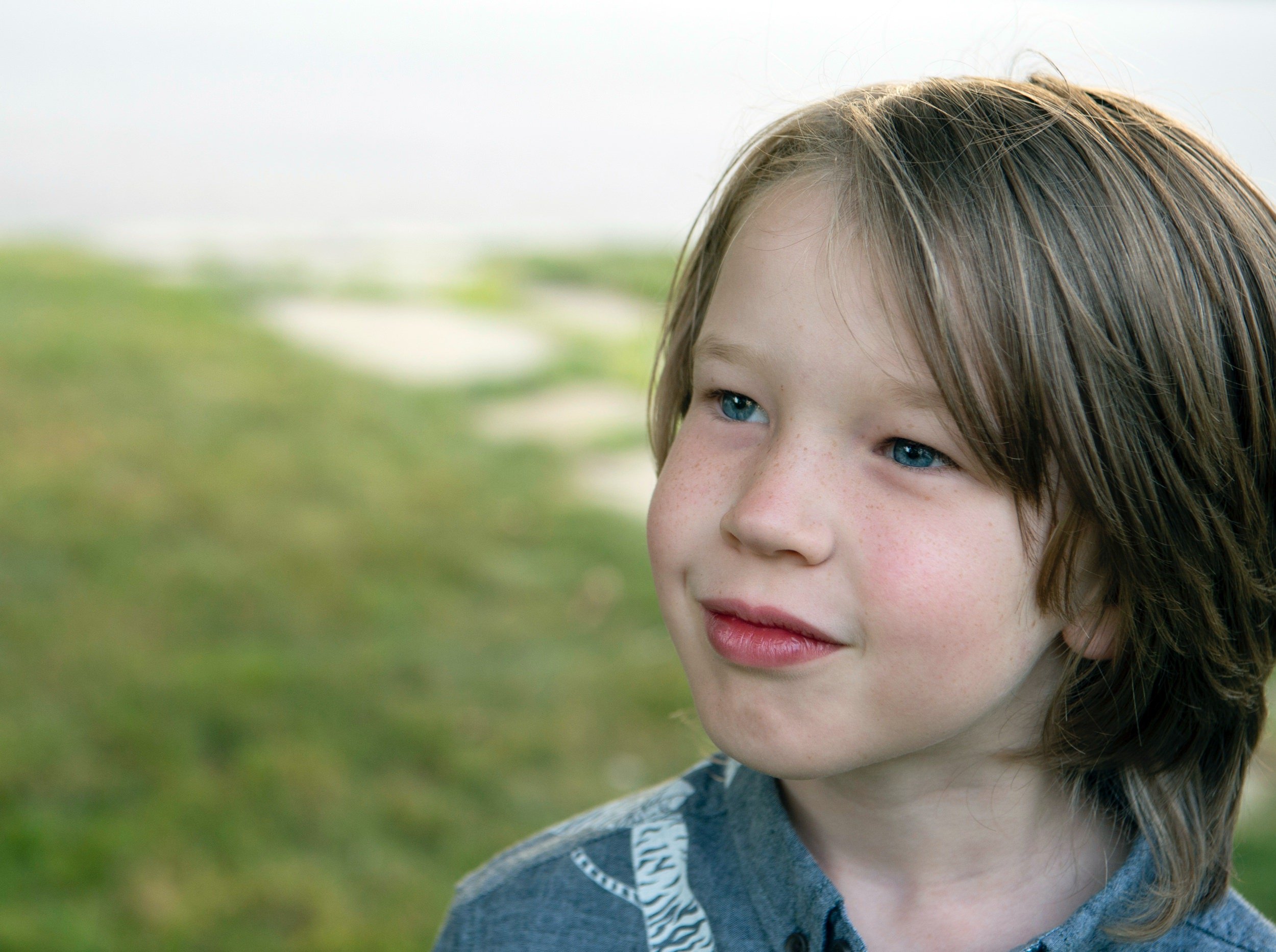 Close-up of a young boy with blue eyes and light brown hair outdoors near a grassy area.
