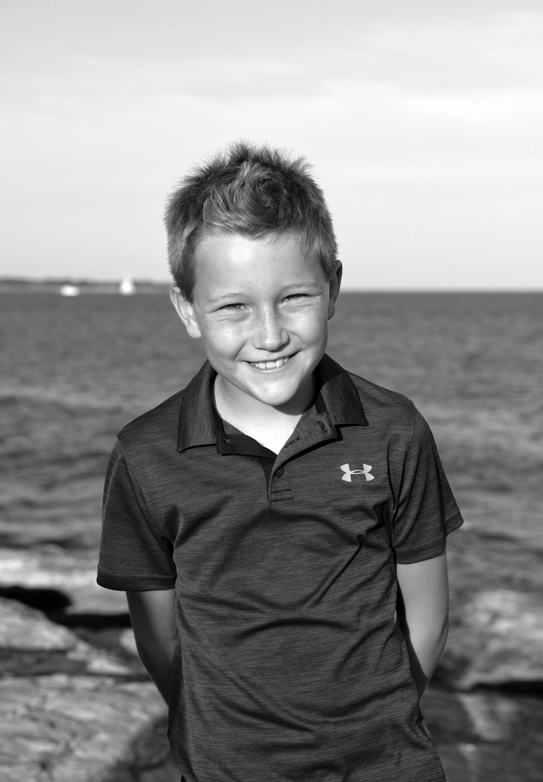 A young boy with short hair smiling at the camera on a beach with water and distant sailboats in the background.