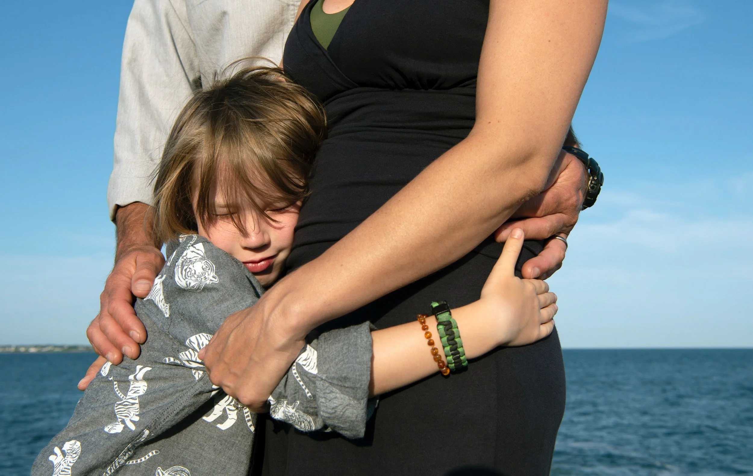 A young boy hugging his mom and dad at Beavertail State Park, Jamestown, Rhode Island. The background features a clear blue sky and water.