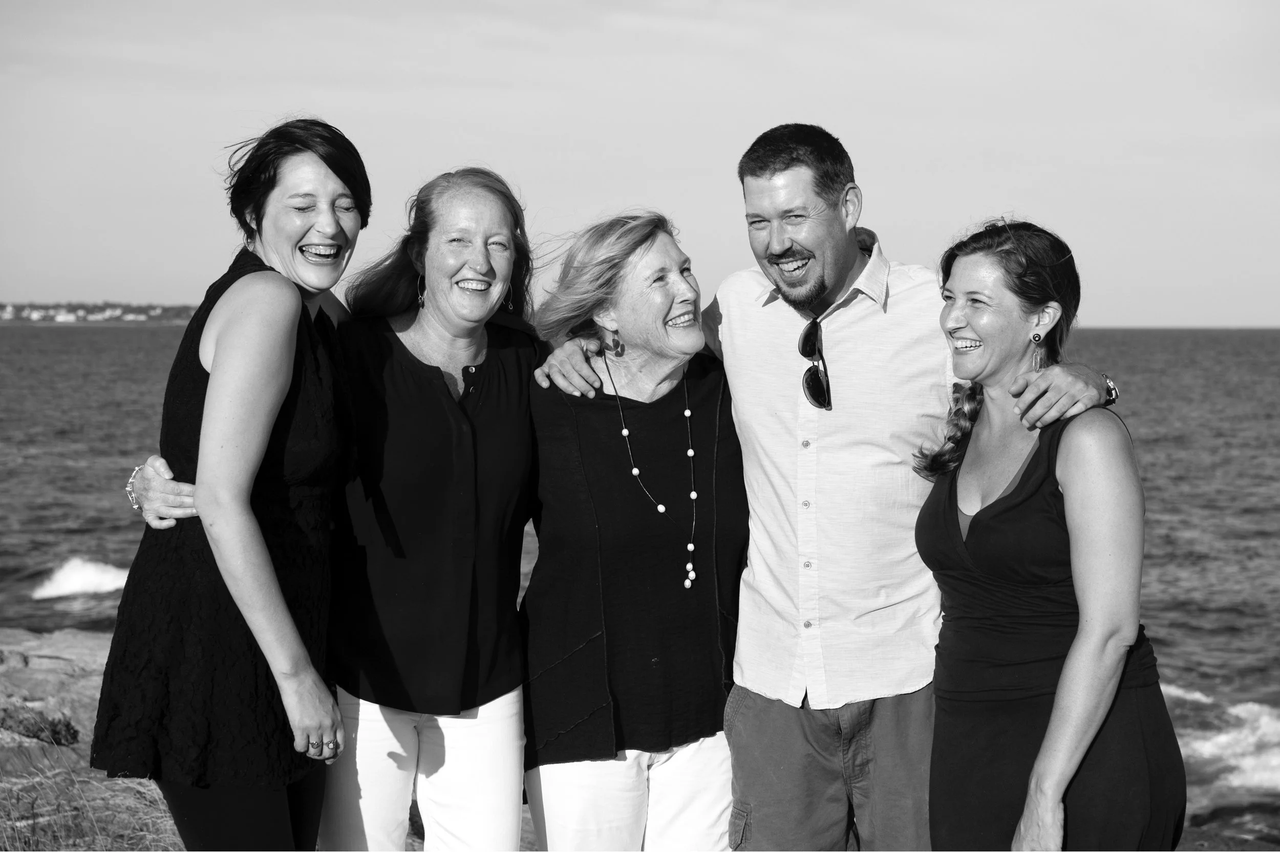 A group of six people, five women and one man, standing close together at the beach, smiling and laughing, in a black-and-white photo.