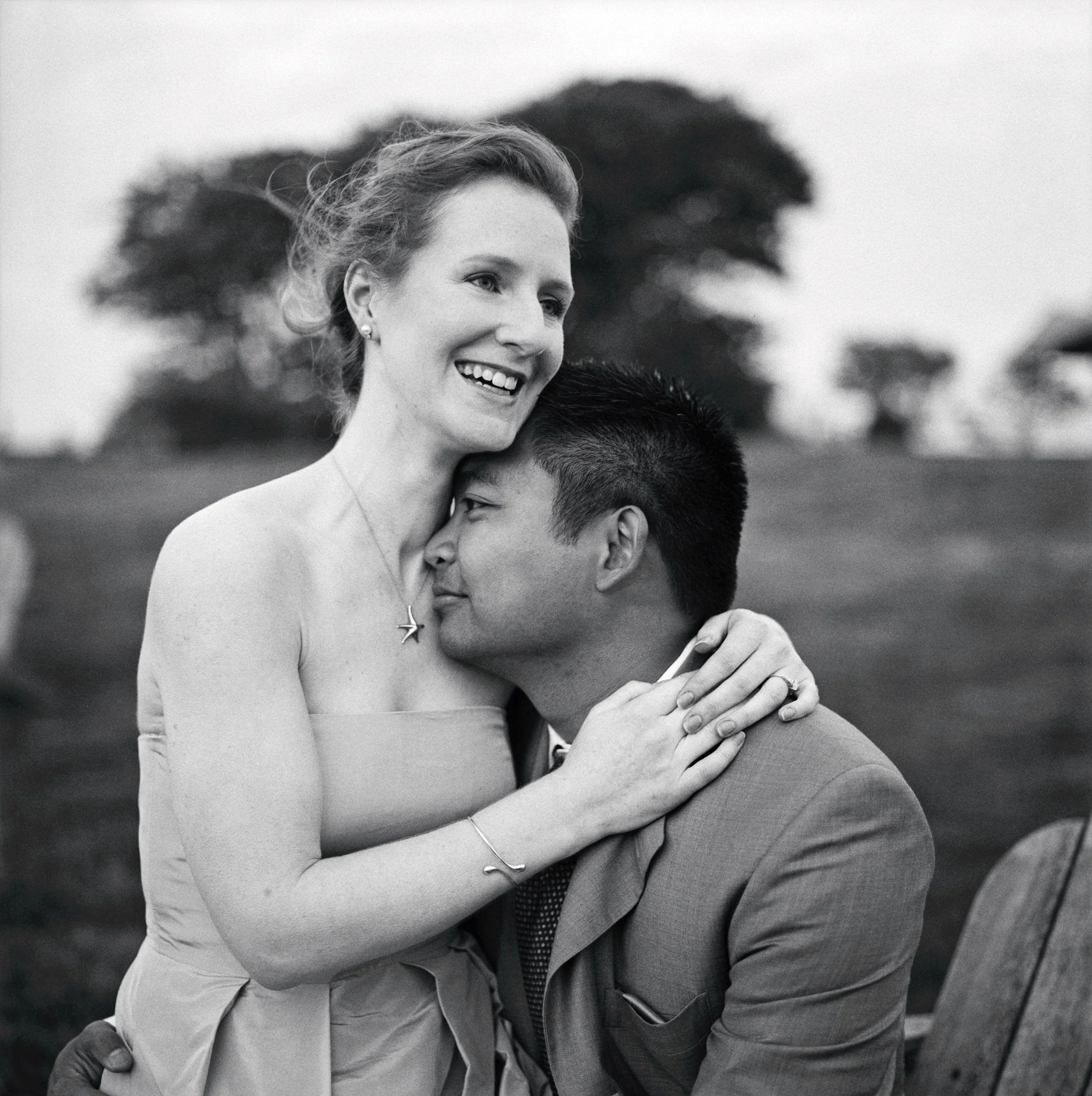 Black and white photo of a smiling woman with curly hair embracing a man, outdoor setting with blurred trees in background. Castle Hill Inn, Newport, RI.