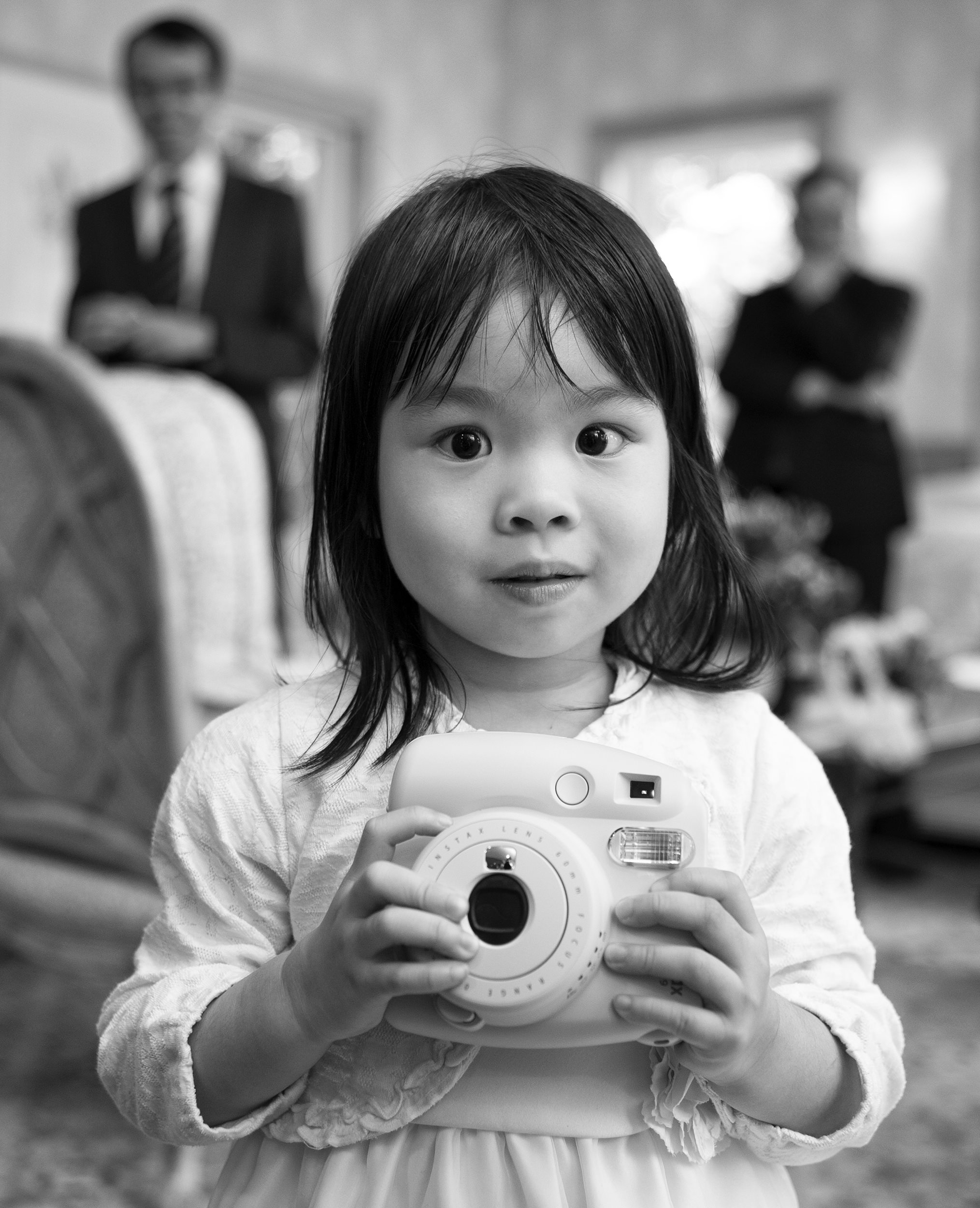 A black and white candid portrait of a young girl surprised at being photographed while holding a camera.