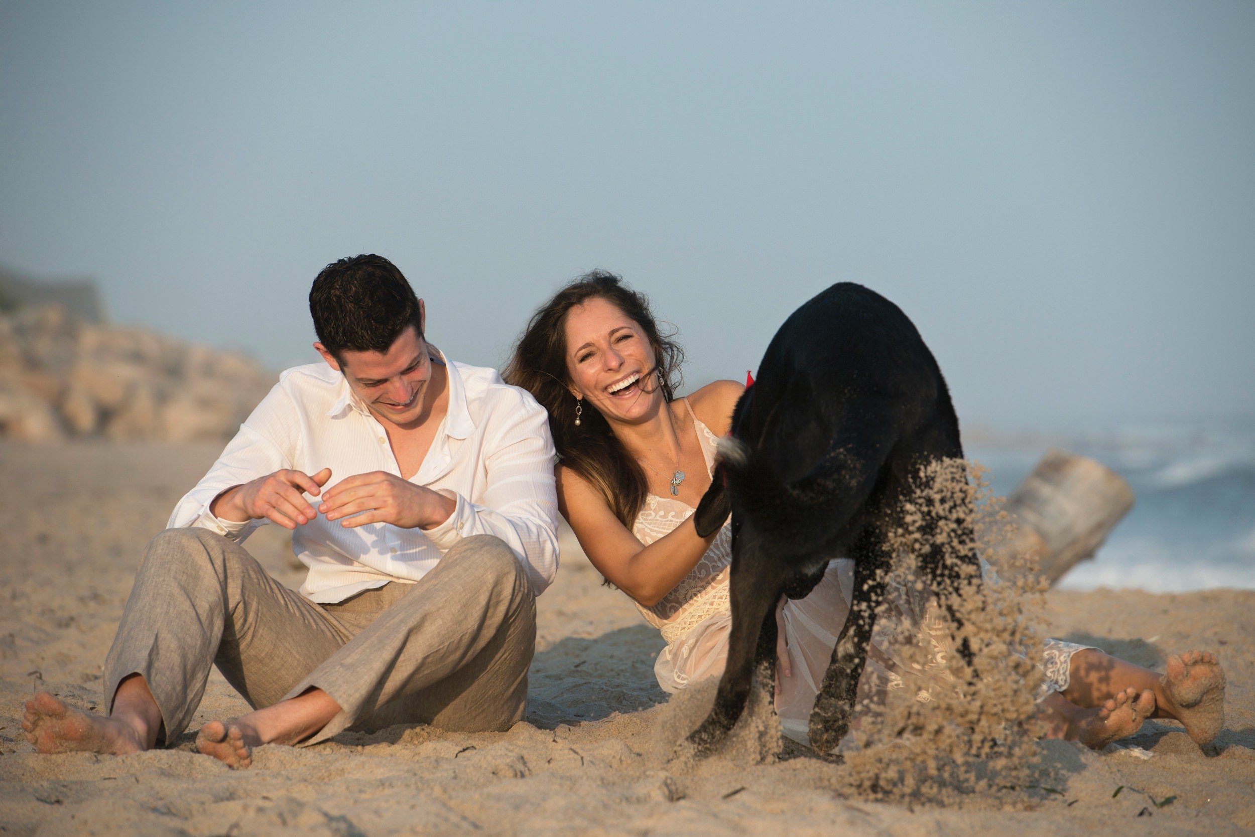 A man and woman sitting on the sandy beach laughing and playing with a black dog, with sand flying in the air, during sunset.