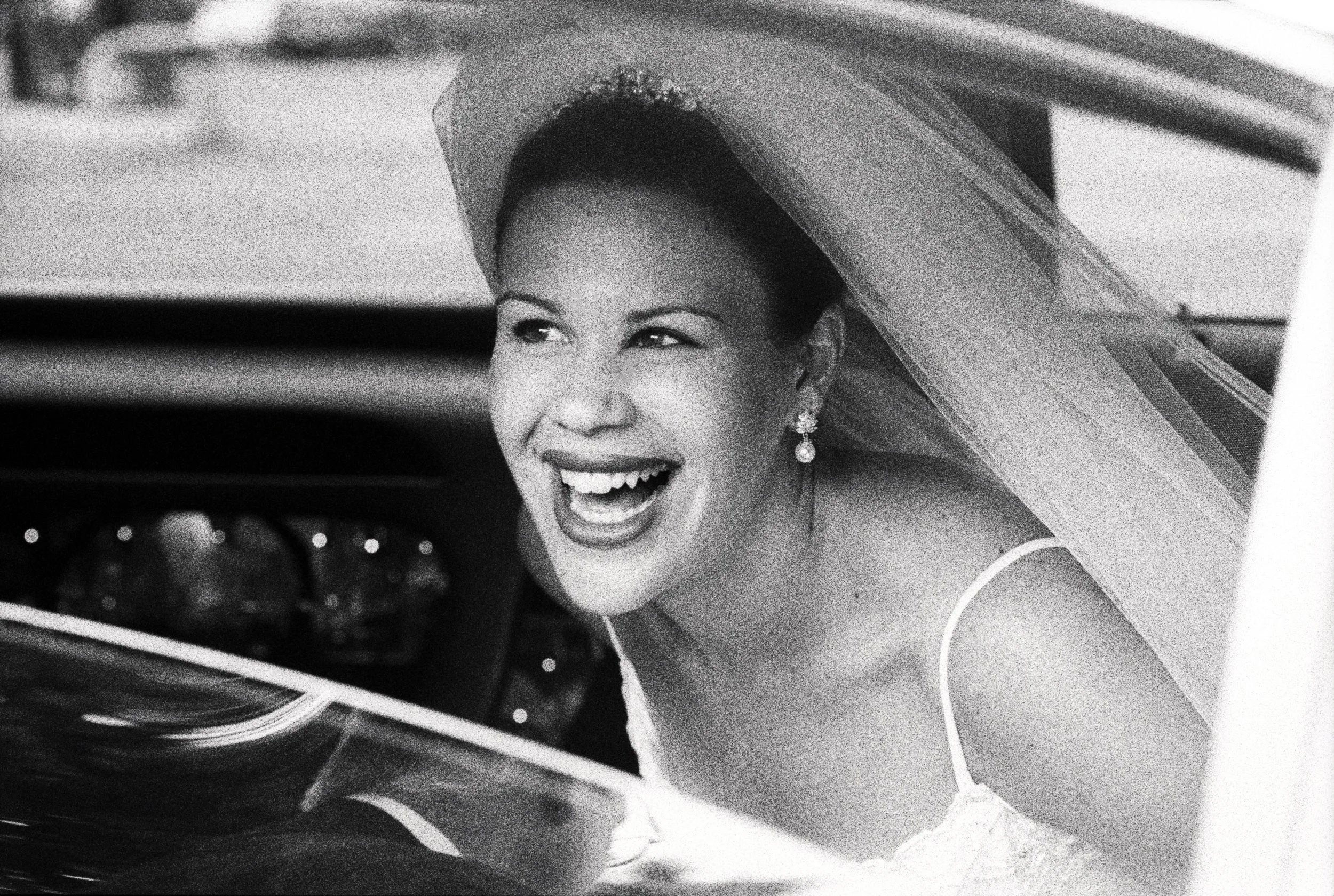 Elegant bride peeks out a car window as she arrives at the ceremony, photographed in grainy black-and-white film.