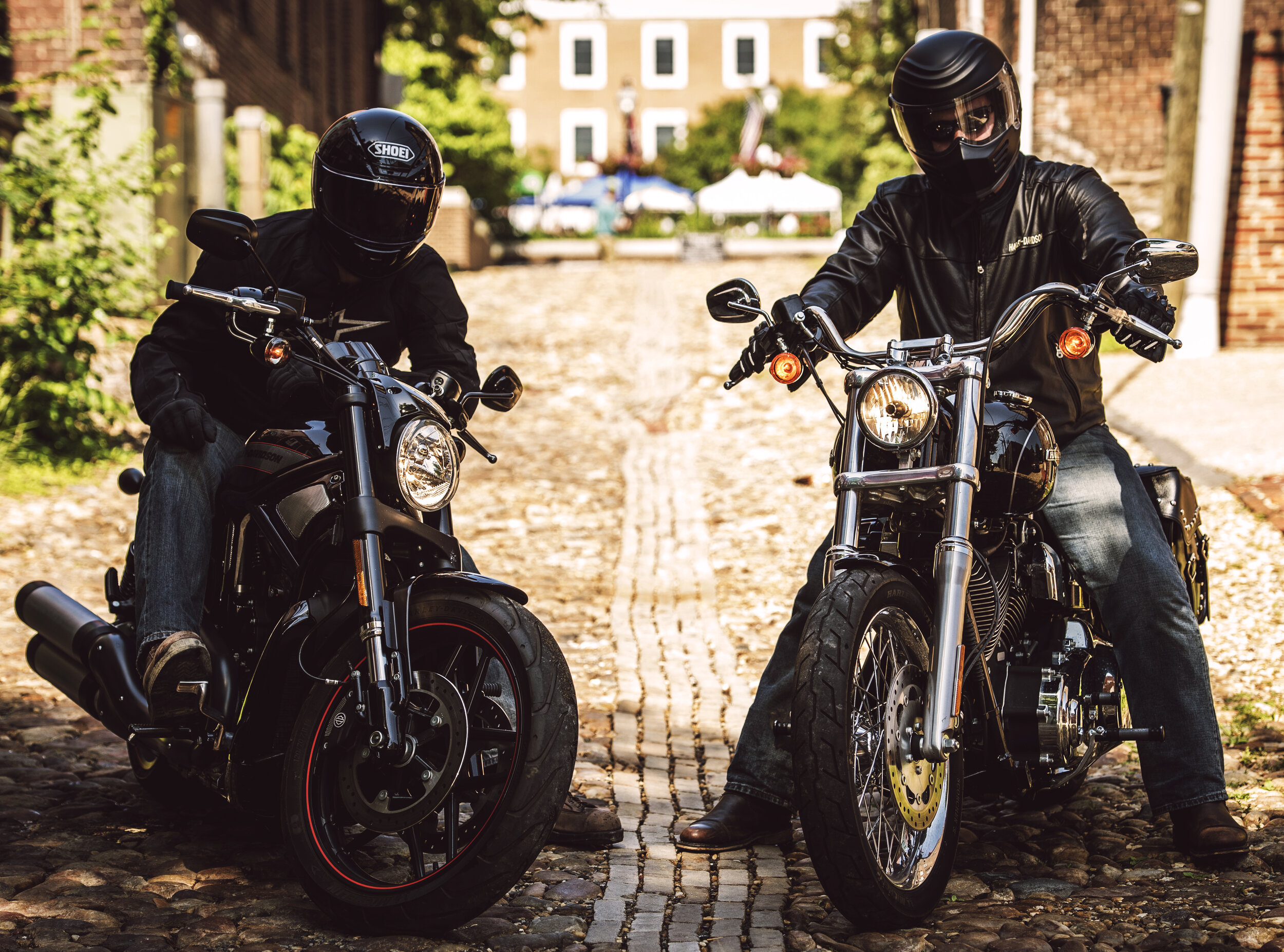 Two motorcyclists wearing black helmets and jackets sitting on black motorcycles on a cobblestone street in a small town, with houses and tents in the background.