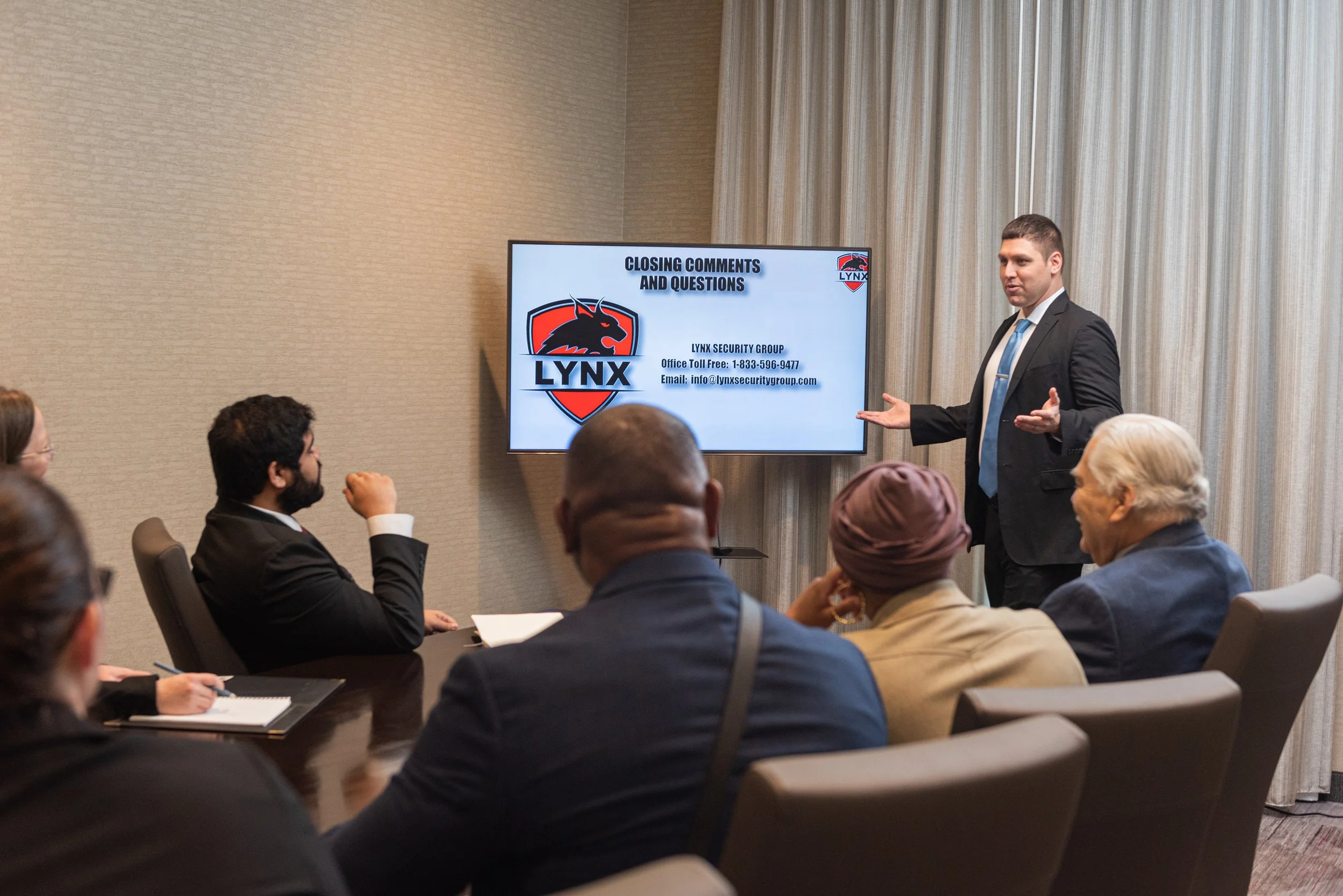 Business presentation in conference room with a man in a gray suit and blue tie speaking to an audience, a screen showing 'Closing Comments and Questions' with Lynx Security Group logo.