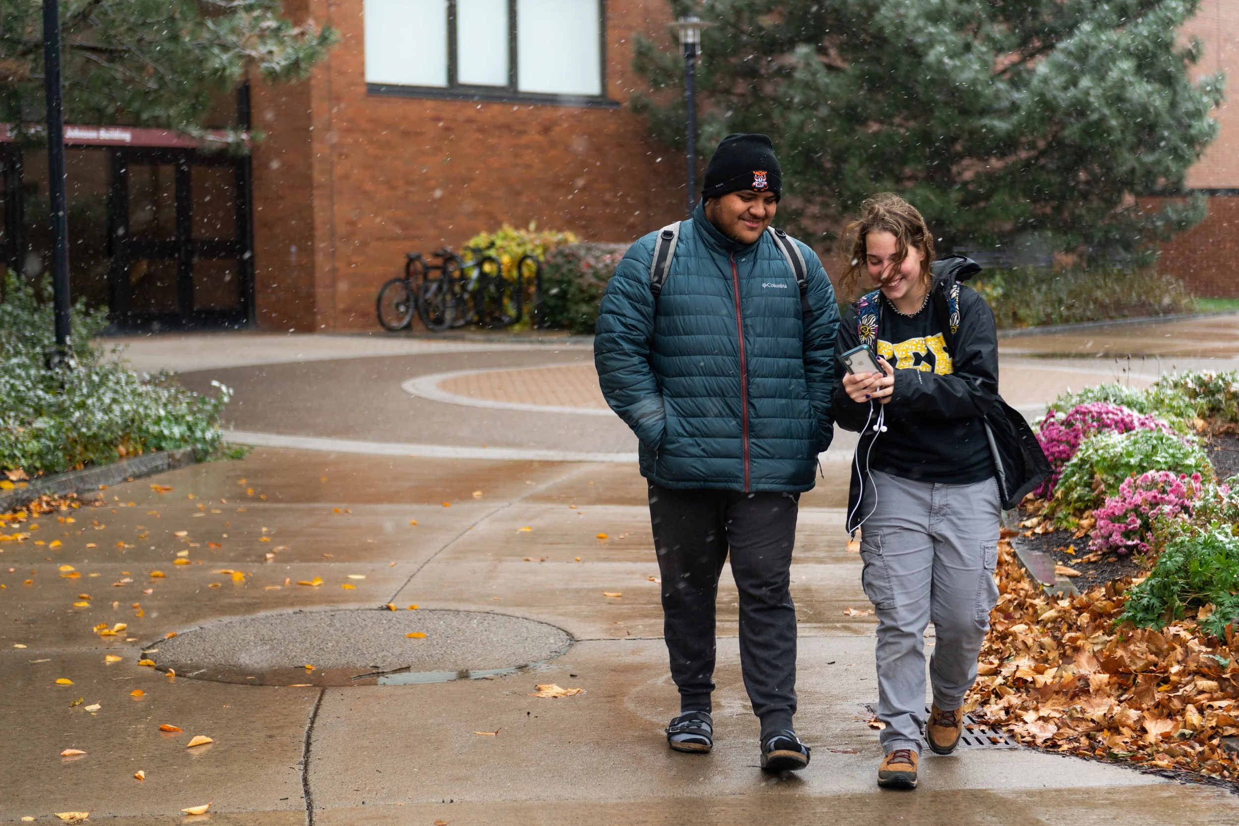 Two young people, a man and a woman, walking and talking on a wet sidewalk during autumn, surrounded by fallen leaves and flowers in bloom, with a building and bicycles in the background.