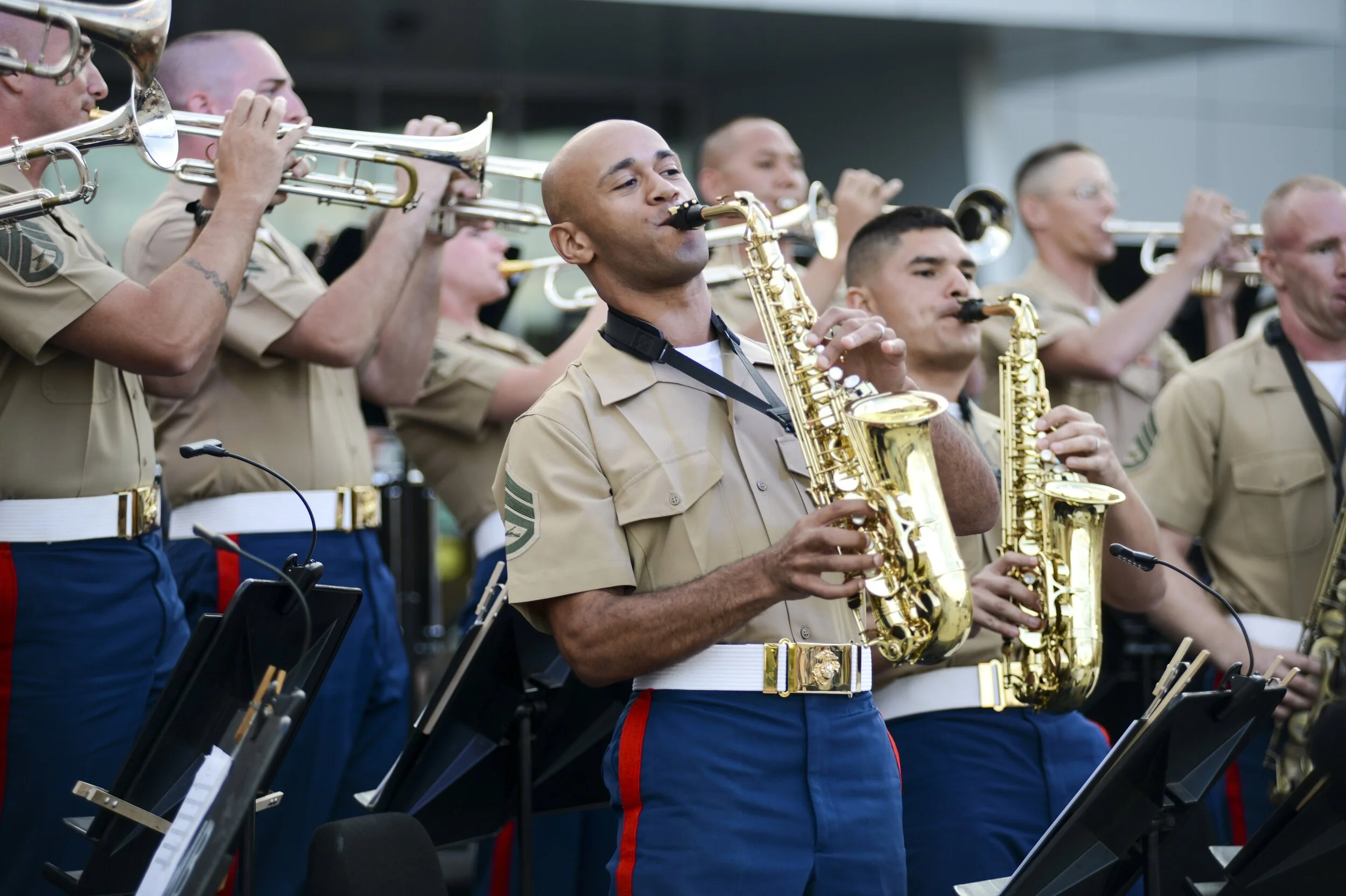 Military band performing, with musicians playing brass and woodwind instruments. The members are dressed in beige shirts with military patches and dark blue pants with red and white stripes.