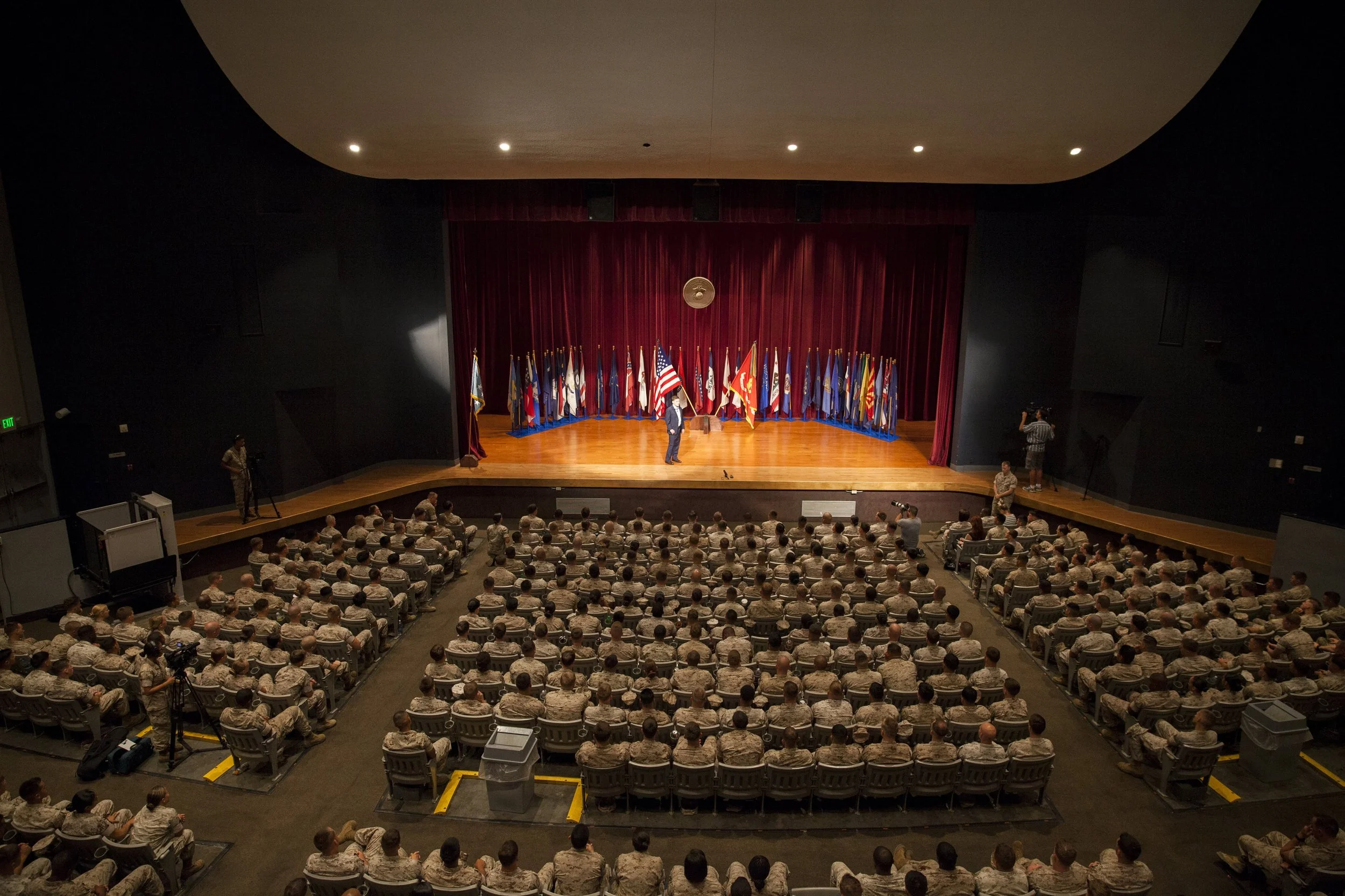 Military personnel attending a ceremony or presentation in an auditorium with a stage, flags, and a speaker.