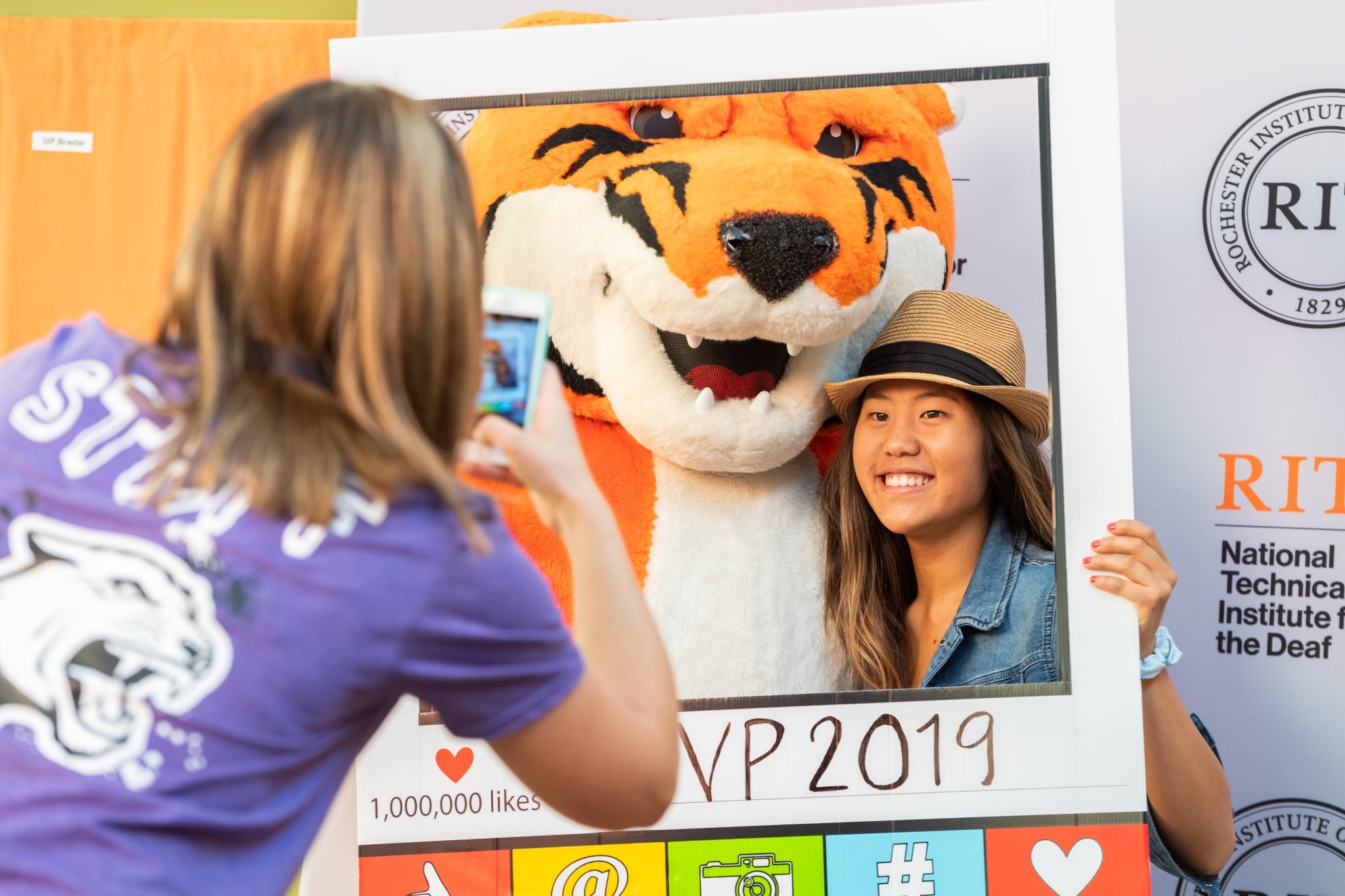A young woman takes a photo of a person in a tiger costume posing behind a large social media celebration board at the Rochester Institute of Technology. The board displays a goal of 1 million likes and various social media icons.