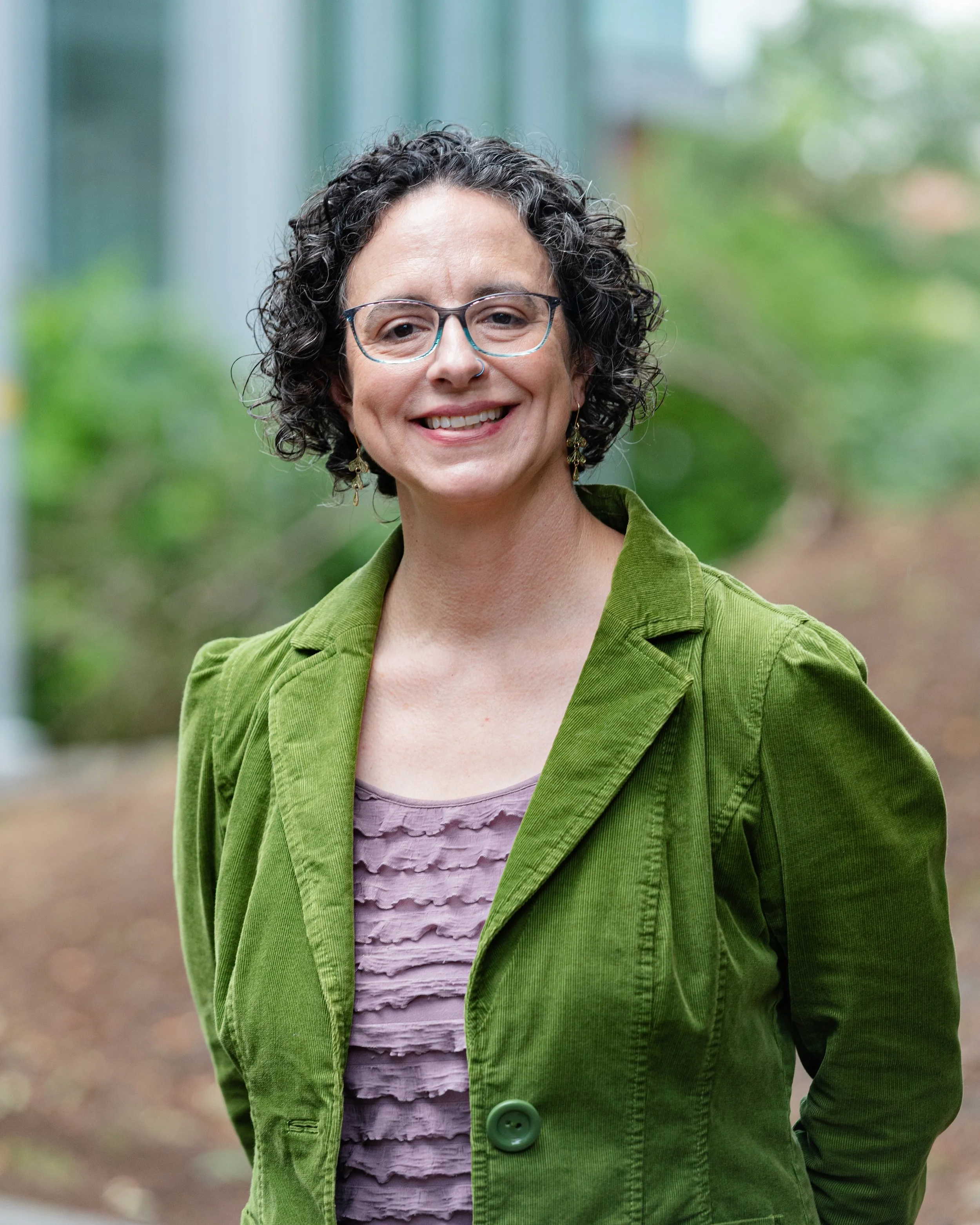 Professional portrait of A smiling woman with glasses and curly dark hair, wearing a green jacket, standing outside a professional office building in Greenbelt Maryland