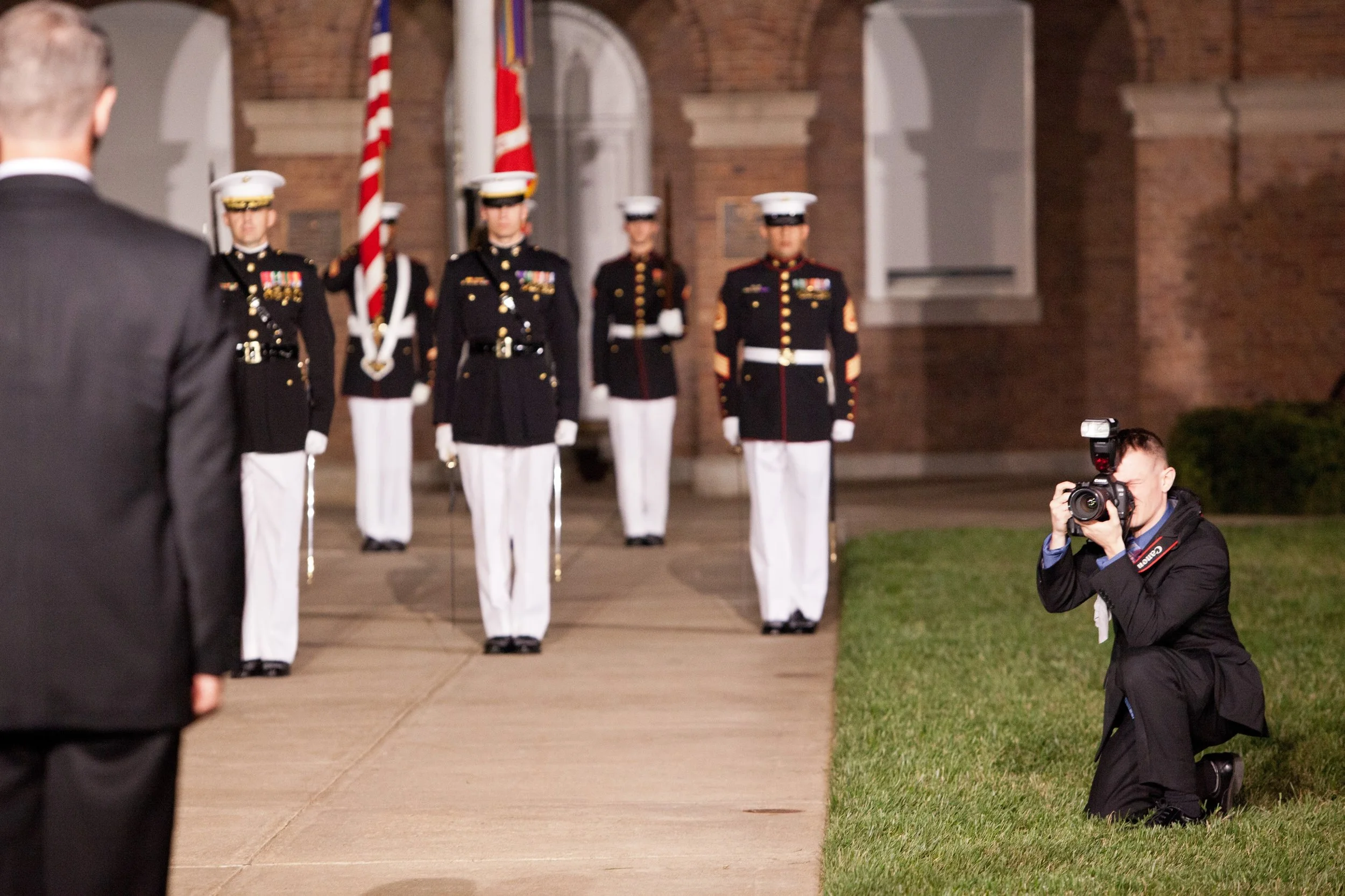 Military guard formation with five soldiers standing at attention in uniform, with a person in a suit in the foreground and a photographer kneeling taking their picture. The background features brick walls and flags.