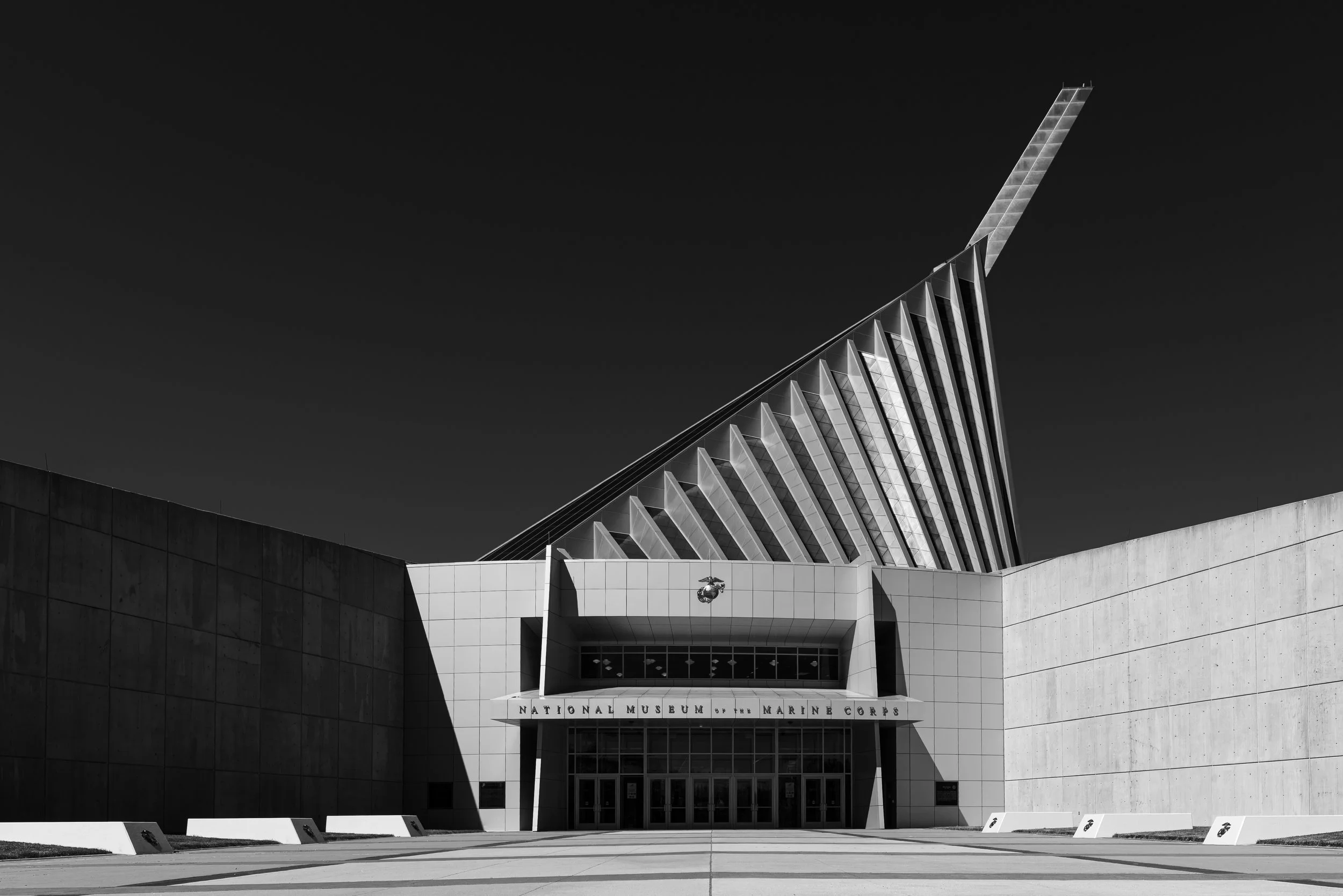 Black and white photo of the National Museum of the Marine Corps building with modern architecture and geometric design, featuring sharp angles and a clear sky background.
