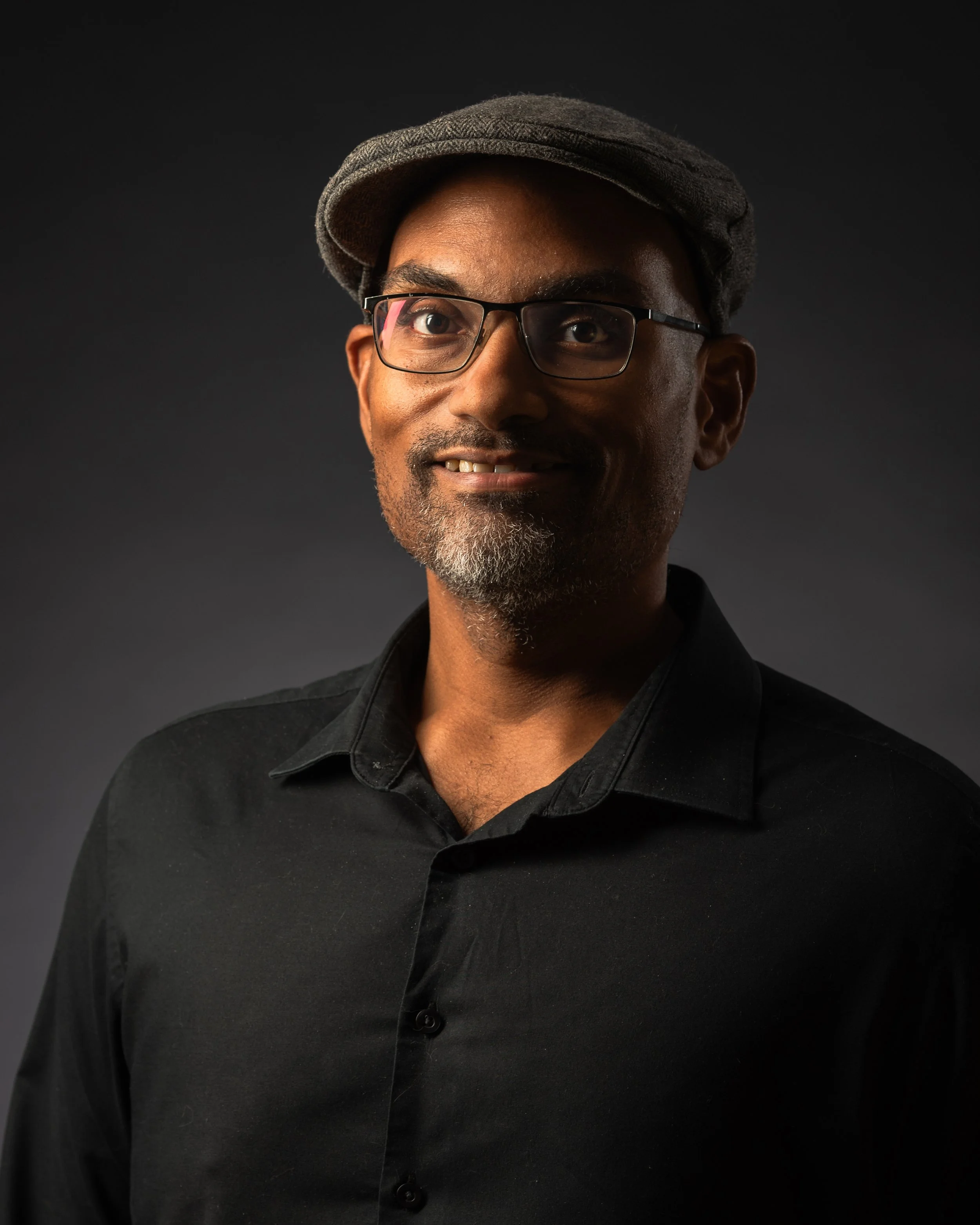 Portrait of a smiling man with glasses, a beard, wearing a dark shirt and a flat cap, against a dark background in a studio setting, Maryland