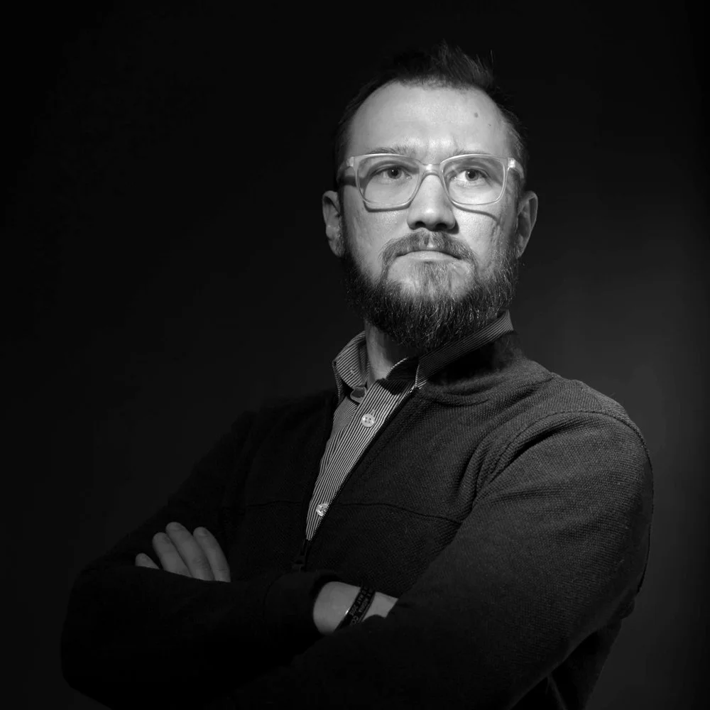A black and white portrait of a man with glasses, a beard, wearing a shirt and sweater, with arms crossed and looking to the side against a dark background.