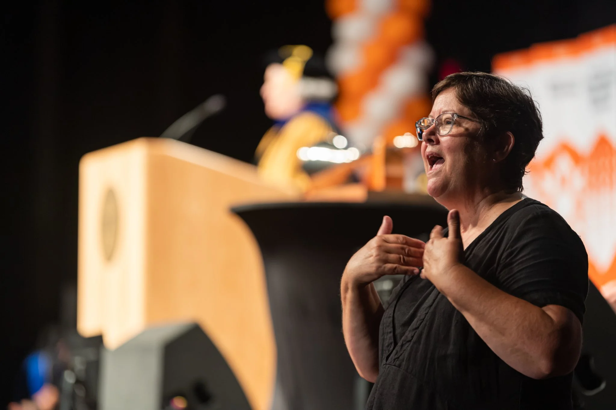 A woman with short brown hair, glasses, and wearing a black top, speaking passionately at a podium during an event, with an out-of-focus figure and orange-themed decorations in the background.
