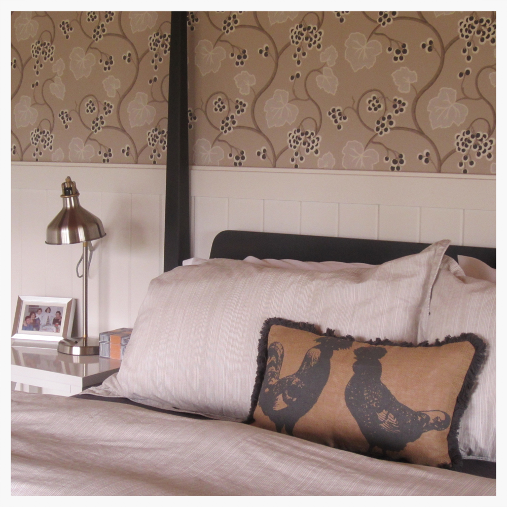 Wine Country retreat master bedroom gets comfy four-post bed in charcoal gray. Walls are covered in neutral-color grapevine motif wallpaper imported from Britain. Extra-tall white wainscoting is a country-modern touch.