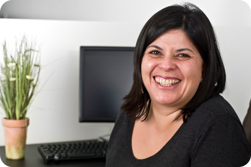 Women cheerfully smiling at the camera while sitting on a desk with a plant and monitor in the background.