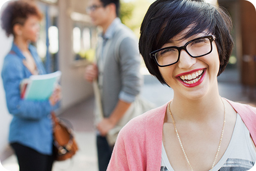 Smiling woman in glass and red lipstick outside posing for the camera. There are two blurred images of individuals (man and woman) speaking to one another . The woman his holding a few books and a book bag.