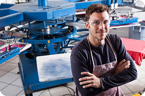 Man wearing eye protectors and gear with his hands crossed in front of him as he smiles at the camera. Machines are in the background.
