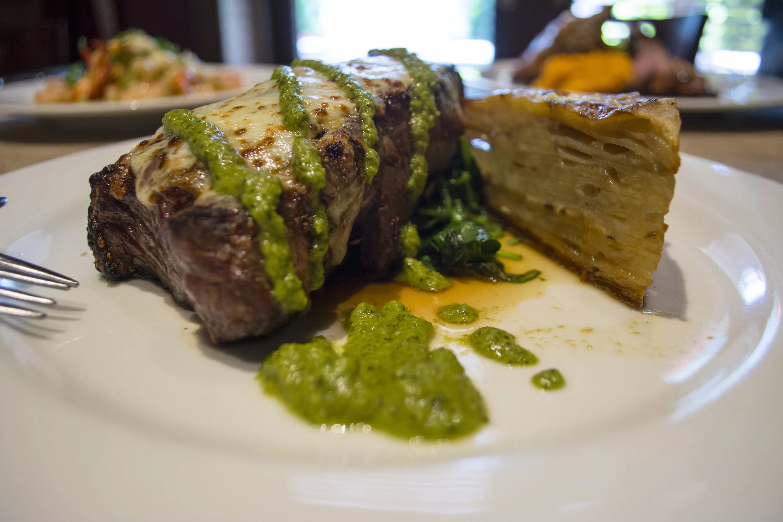 The creative, yet classic Dry Aged NY Strip steak is topped with cheese and chimichurri sauce paired with a side of potato au gratin and sauteed spinach.