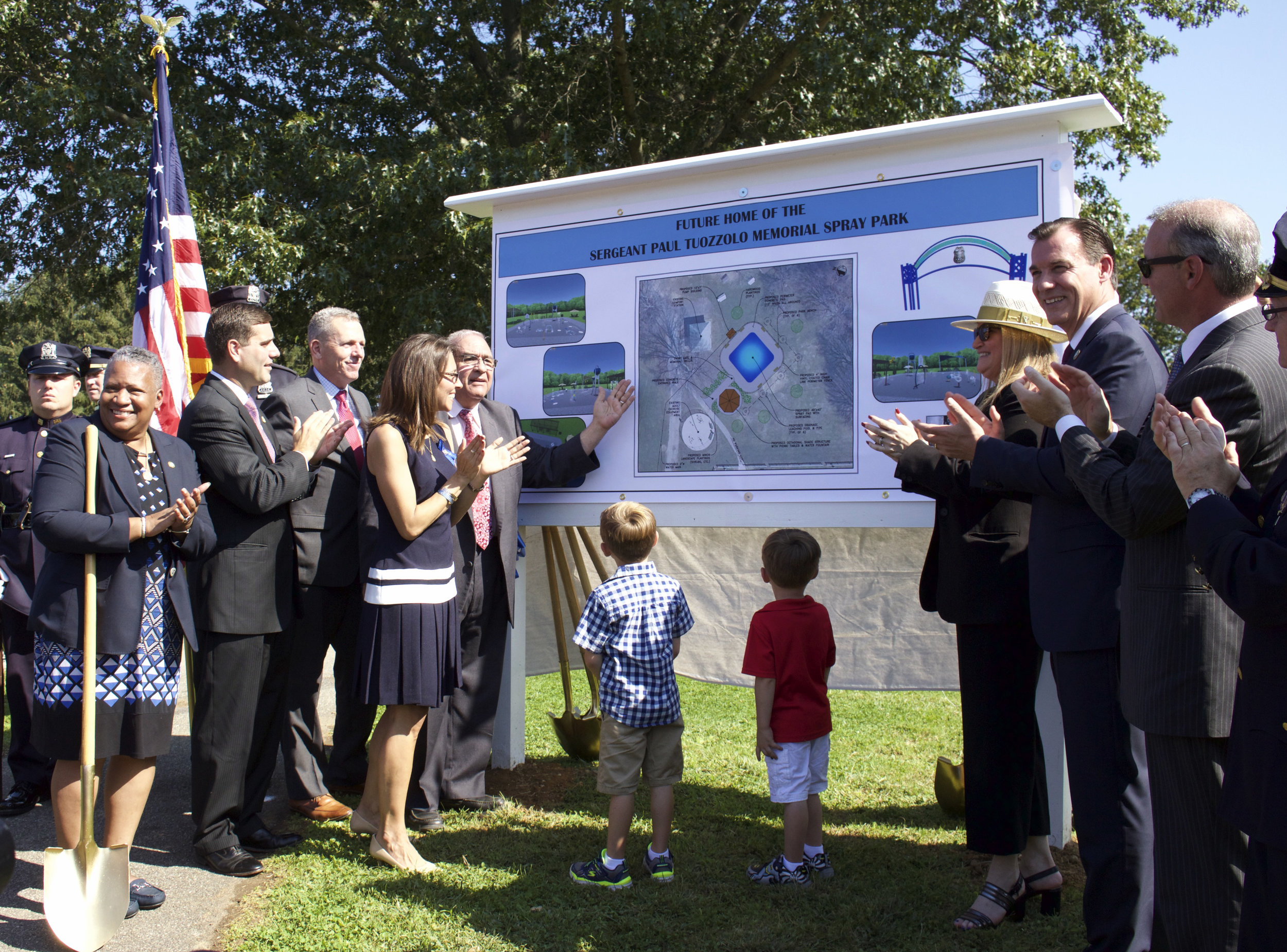   Austin, 5, and Joseph, 4, Tuozzolo, center, look up at the sign depicting plans for the Sgt. Paul Tuozzolo Memorial Spray Park, set to open in Elwood Park next summer.   Long Islander News photo/Janee Law  