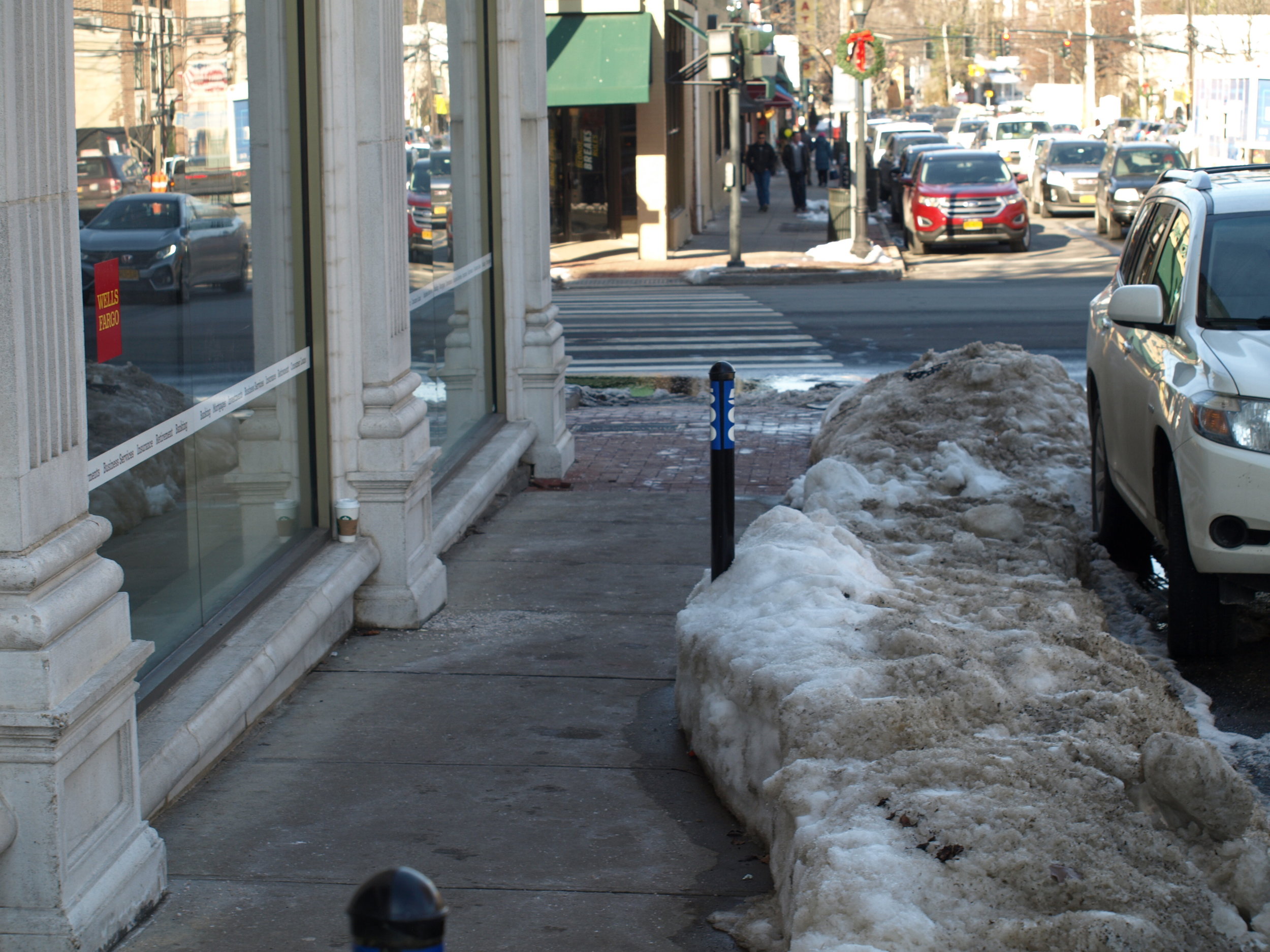   Left, the western sidewalk along Wall Street near the intersection with Main Street in Huntington village is pictured Wednesday after Sunday night’s snow cleanup operation. Right, Wall Street is covered in snow by Winter Storm Grayson last Thursday