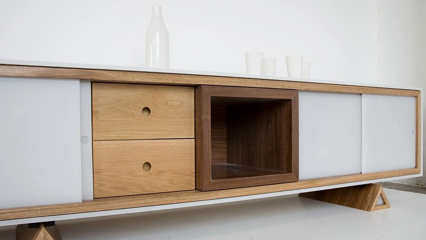 Meuble en bois clair avec deux tiroirs, un espace ouvert et deux portes en façade, posé sur des pied en bois inclinés, décoration minimaliste avec vases et verres blancs sur le dessus.