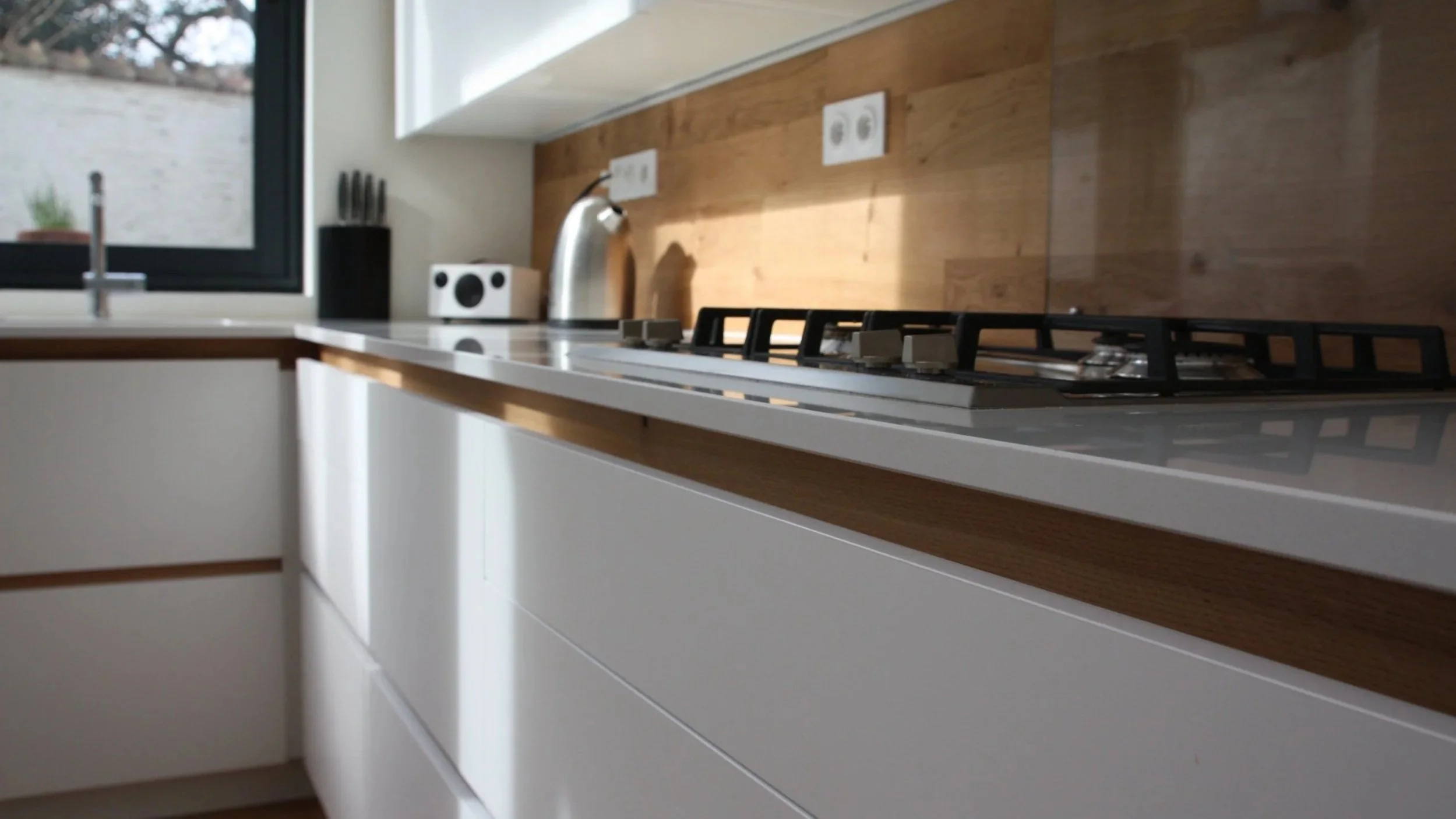 Cuisine moderne avec comptoir blanc, plan de travail en bois, évier, four à gaz noir, fenêtre, et prises électriques.