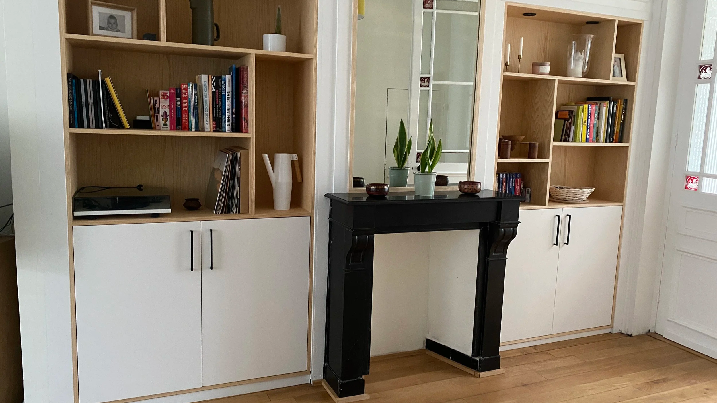 Étagères en bois avec livres, objets décoratifs et plantes, cheminée noire au centre, sol en bois, porte fenêtre en arrière-plan.