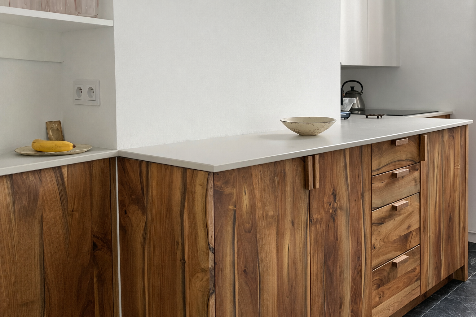 Une cuisine avec des meubles en bois et un plan de travail blanc, quelques objets dont une banane, un bol et une bouilloire visible.