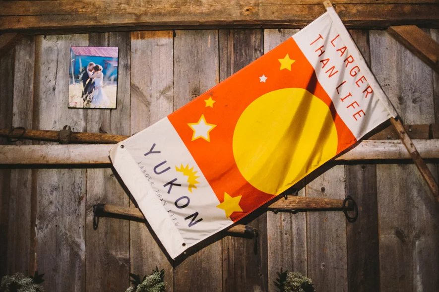 A flag with red, yellow, and white colors, displaying the words "Larger Than Life" and "Yukon" along with a large yellow circle and yellow stars, mounted on a wooden wall.