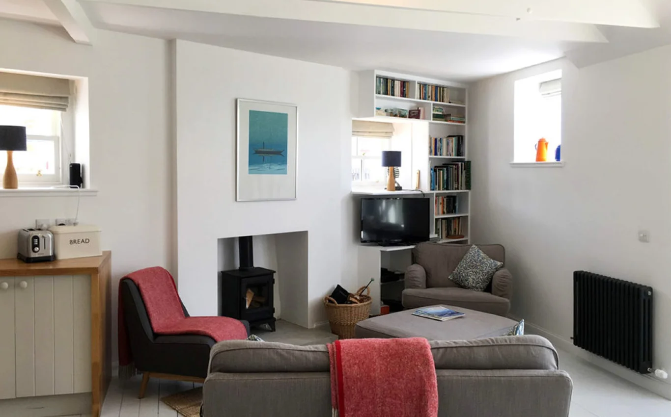 Working within the footprint of the existing cottage challenged us to optimise all available space, minimising the floor areas dedicated to circulation in order to free up more space for living, and tucking carefully-designed storage units within the irregular nooks and crannies. The original floor plan was obstructed by a masonry chimney stack, a later addition to the property which occupied valuable space internally. This was removed and the hearth reinstated in its original location to the centre of the angled gable wall, with a new masonry chimney constructed to the gable wall head as it would have been when the cottage was built. To further augment the open and spacious nature of the new interior the main living space was opened up into the roof volume, with the exposed and white-painted timber structure adding a subtle sculptural quality overhead.