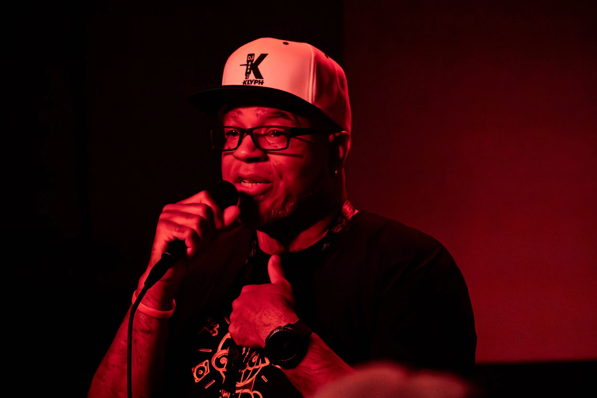 A person with glasses and a cap holding a microphone and speaking, illuminated by red lighting.