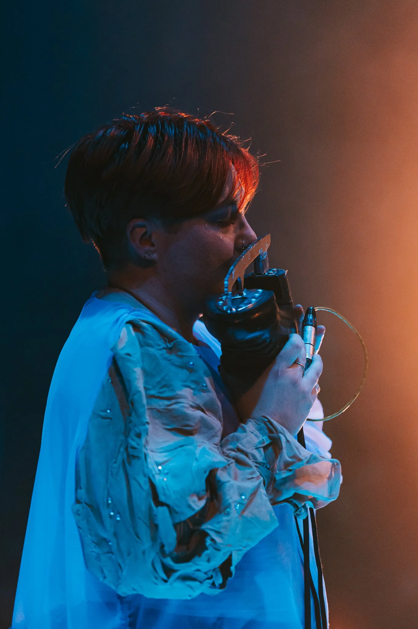 jo, a white non binary person with reddish hair, is holding the blade of a figure skating boot to their mouth. their eyes are closed. they are wearing a blue chiffon shirt with a wrinkled, bedazzled sleeve